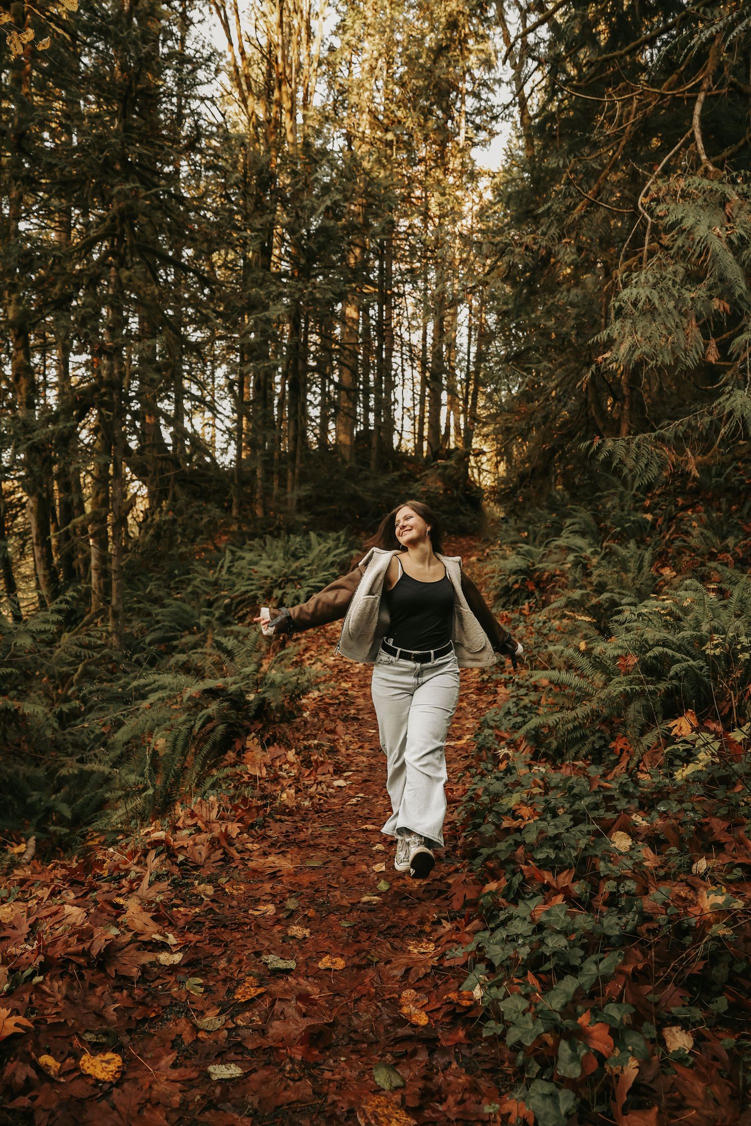 A person walks along an autumn forest trail surrounded by tall evergreen trees and fallen leaves in warm lighting.