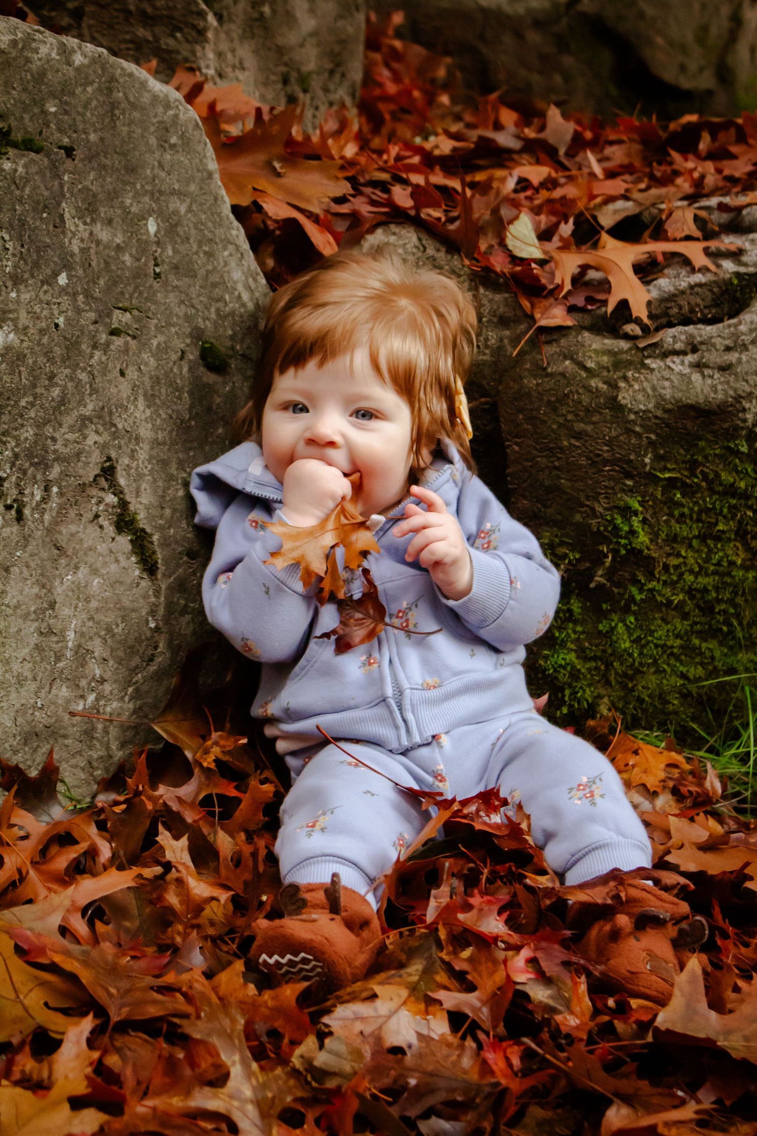 A small figure in light blue sleepwear sits among autumn leaves near moss-covered rocks on a fall day.