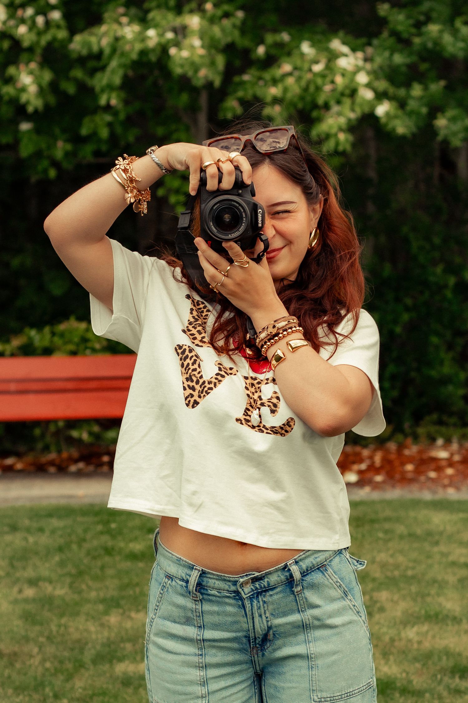 A photographer holds a professional DSLR camera while wearing a white t-shirt and jeans in a park setting.