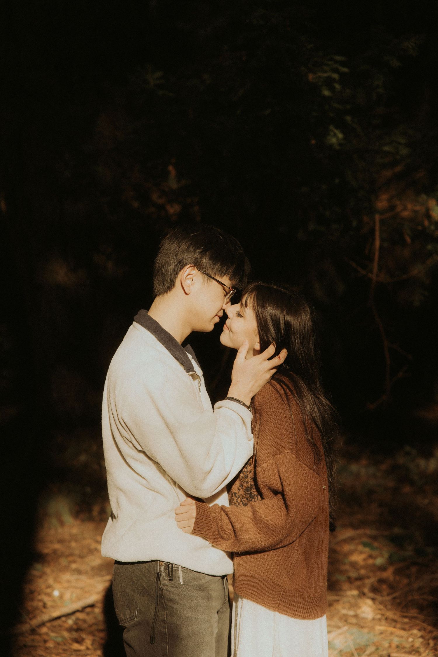 A couple shares an intimate embrace in a dark forest setting illuminated by warm golden light.