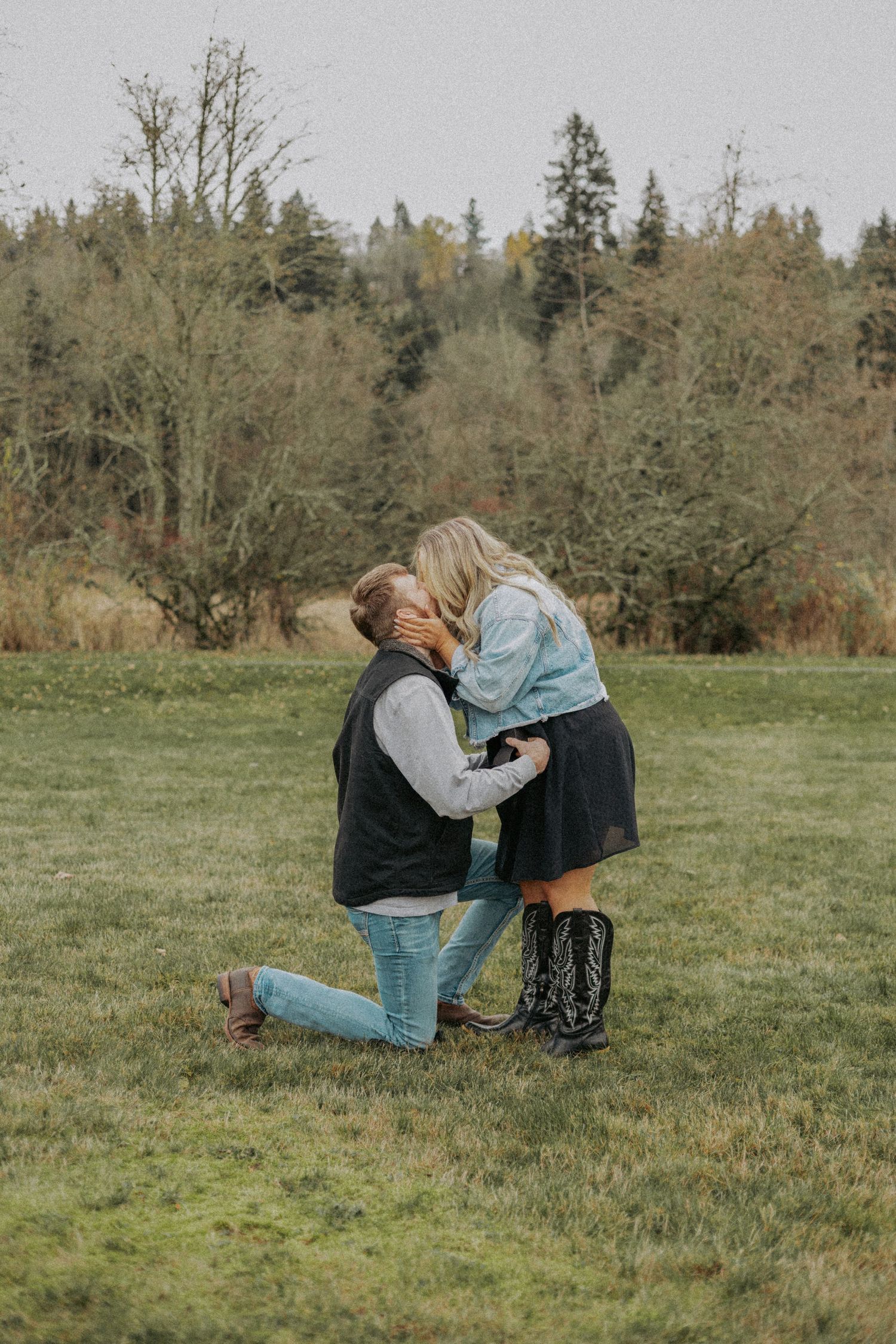 A romantic proposal moment captured in a green field with trees in the background on an overcast day.