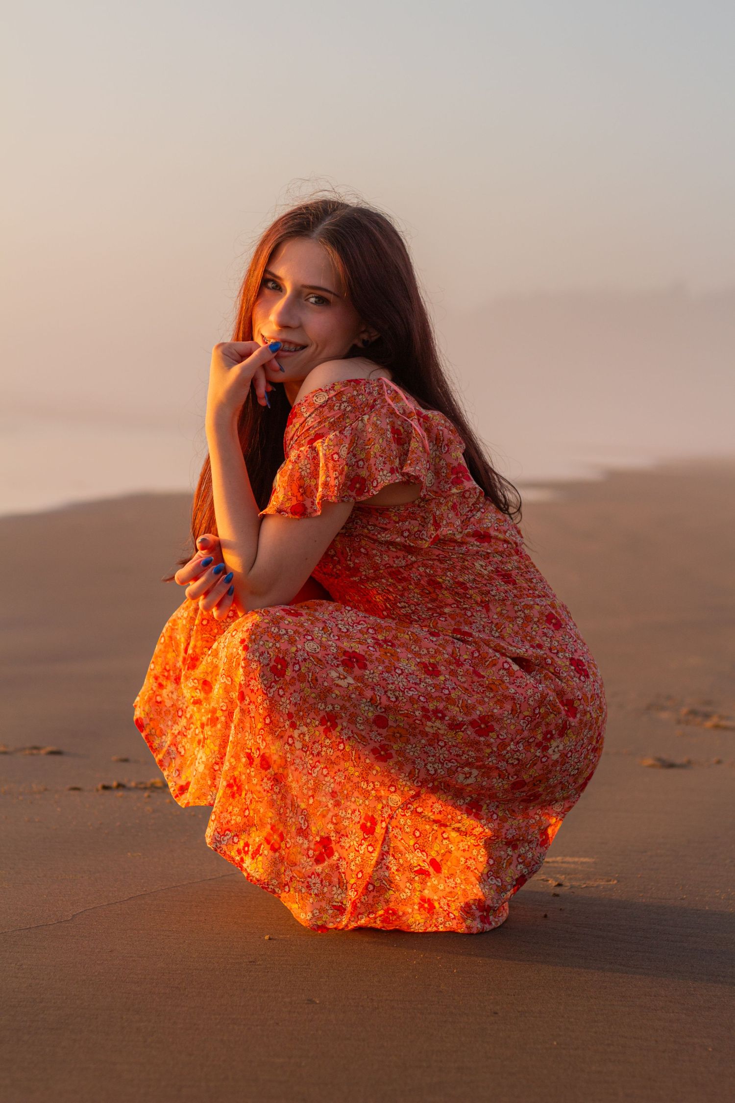 Person in flowing orange floral dress crouches on sandy beach at sunset with warm golden lighting.