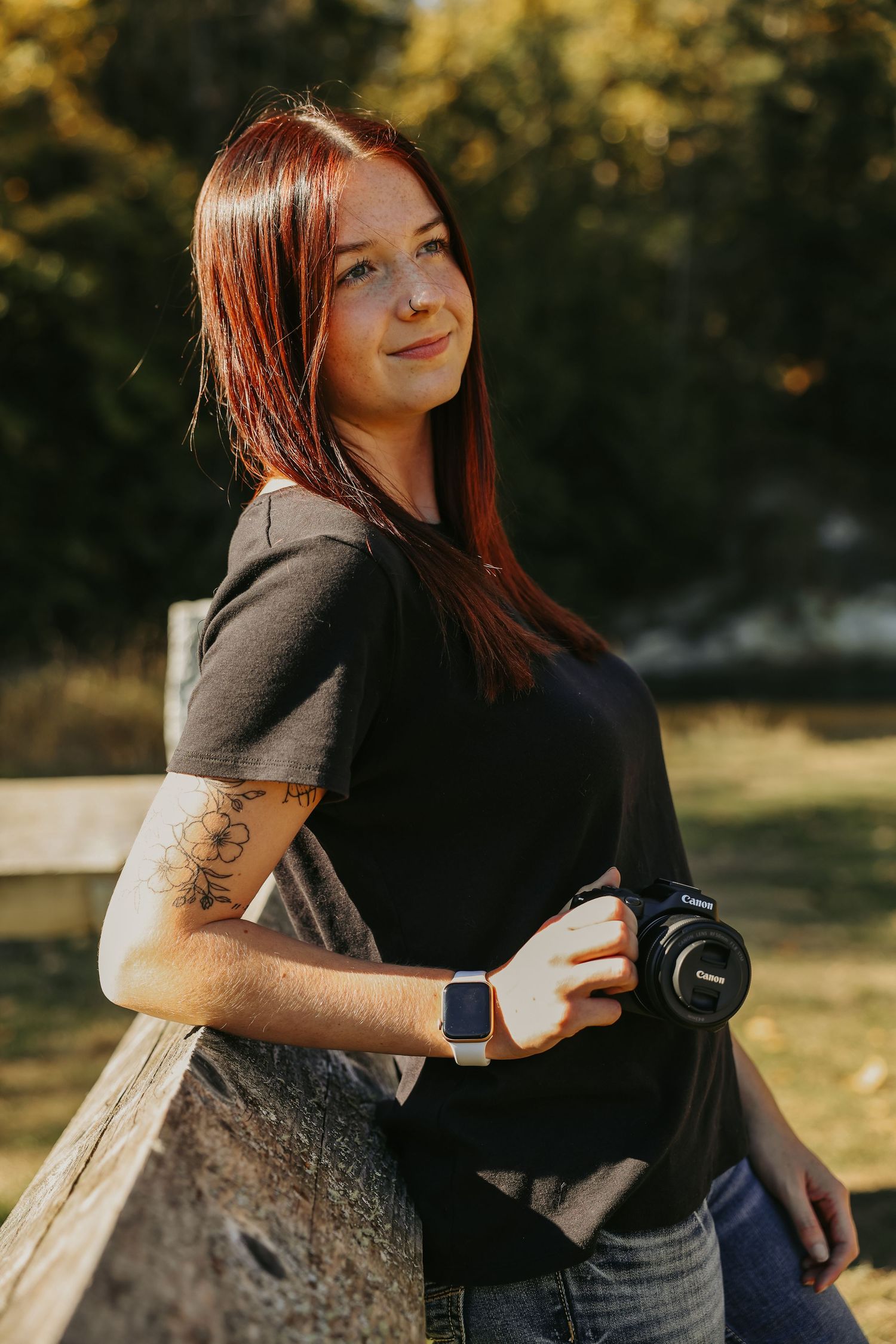 A photographer with red hair holds a camera while sitting outdoors in autumn lighting.