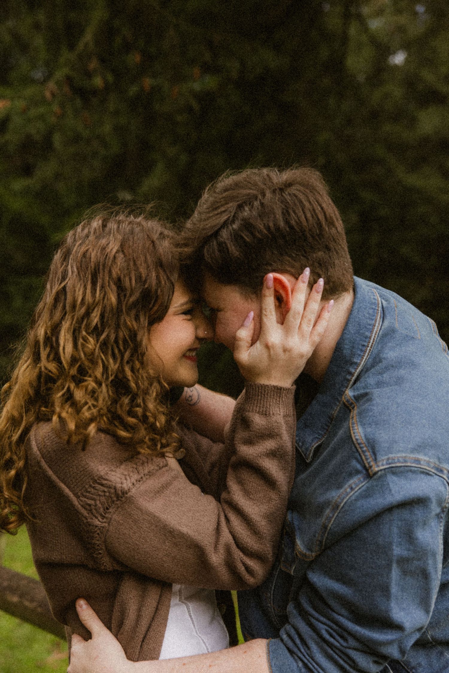 Romantic couple sharing an intimate moment outdoors with foreheads touching while wearing brown sweater and denim jacket.