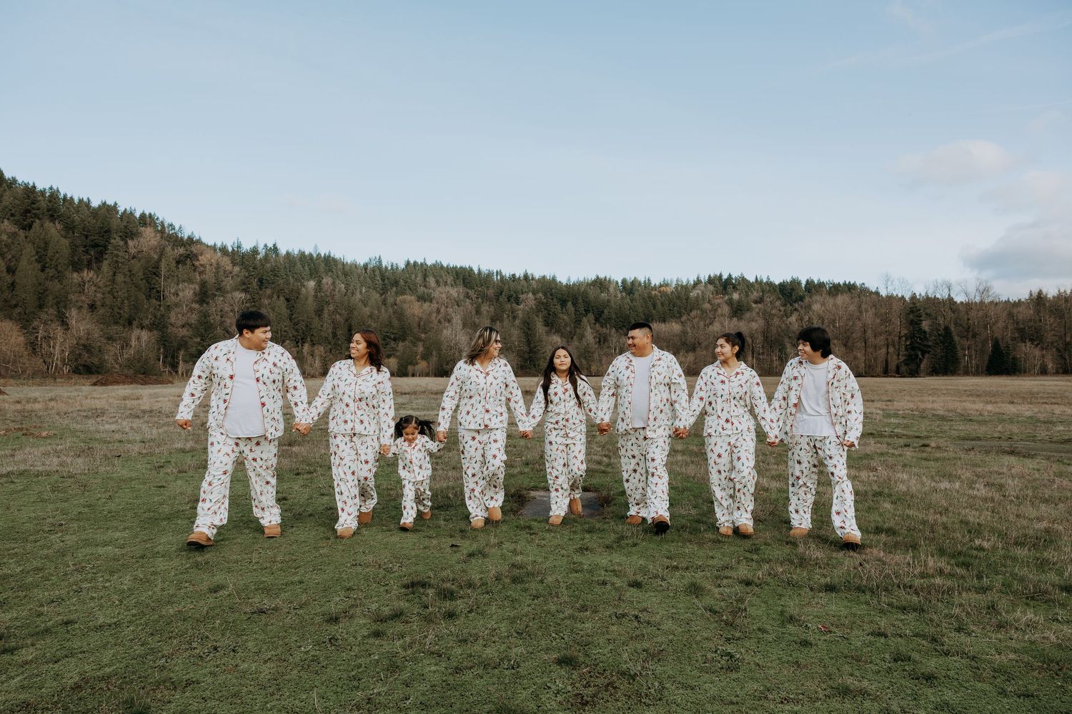 A group of people wearing matching white outfits hold hands while walking in a line across a grassy field with mountains behind.