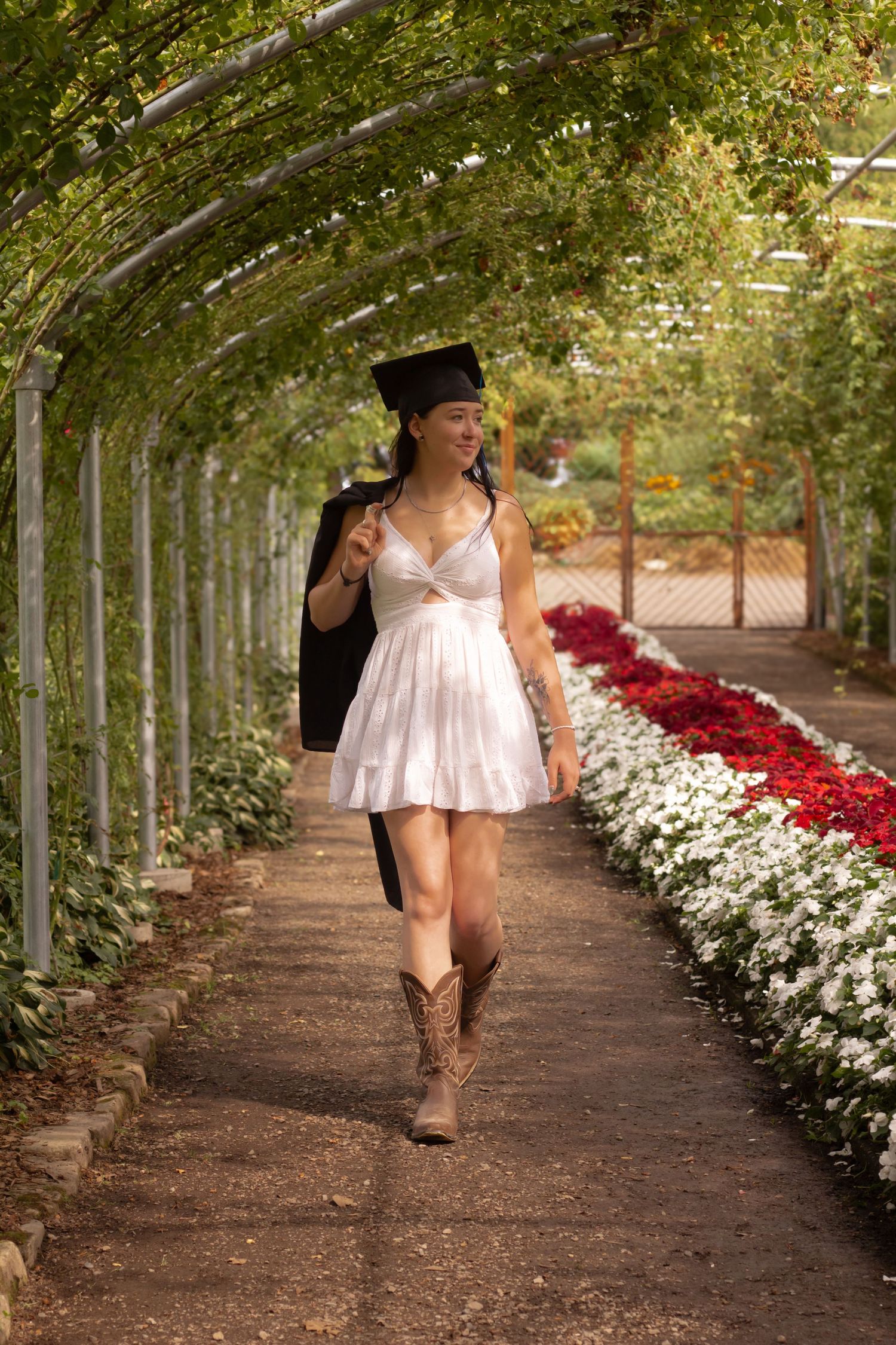 Graduate in a white dress and cowboy boots walking through a garden arch with blooming flowers.