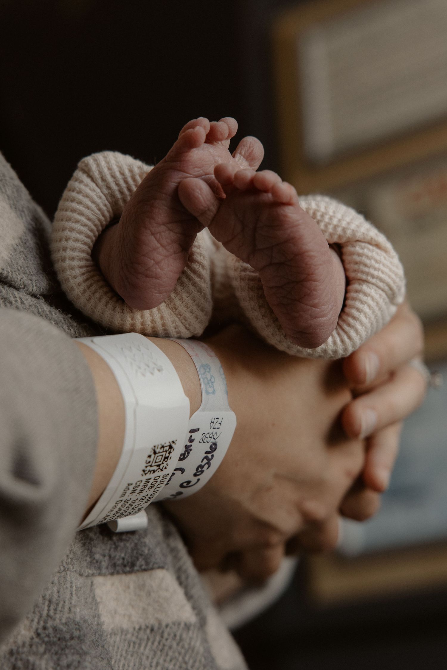 Newborn baby feet held gently by a parent wearing a hospital wristband, wrapped in a cozy knit outfit.