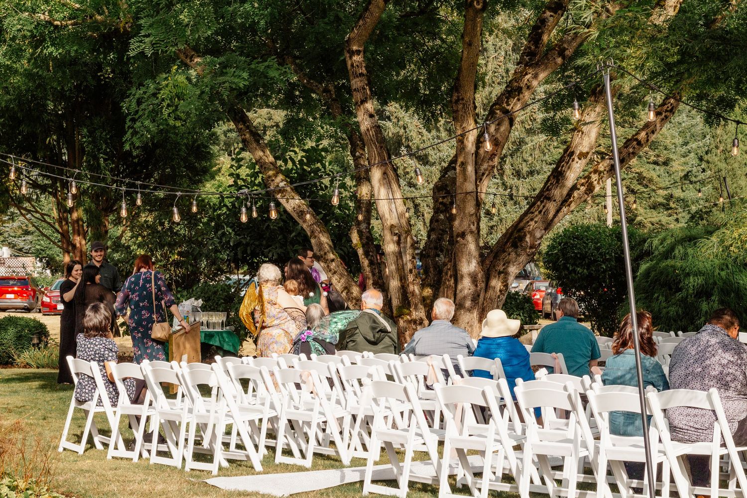 White folding chairs set up for an outdoor ceremony under large trees with guests seated in the background.