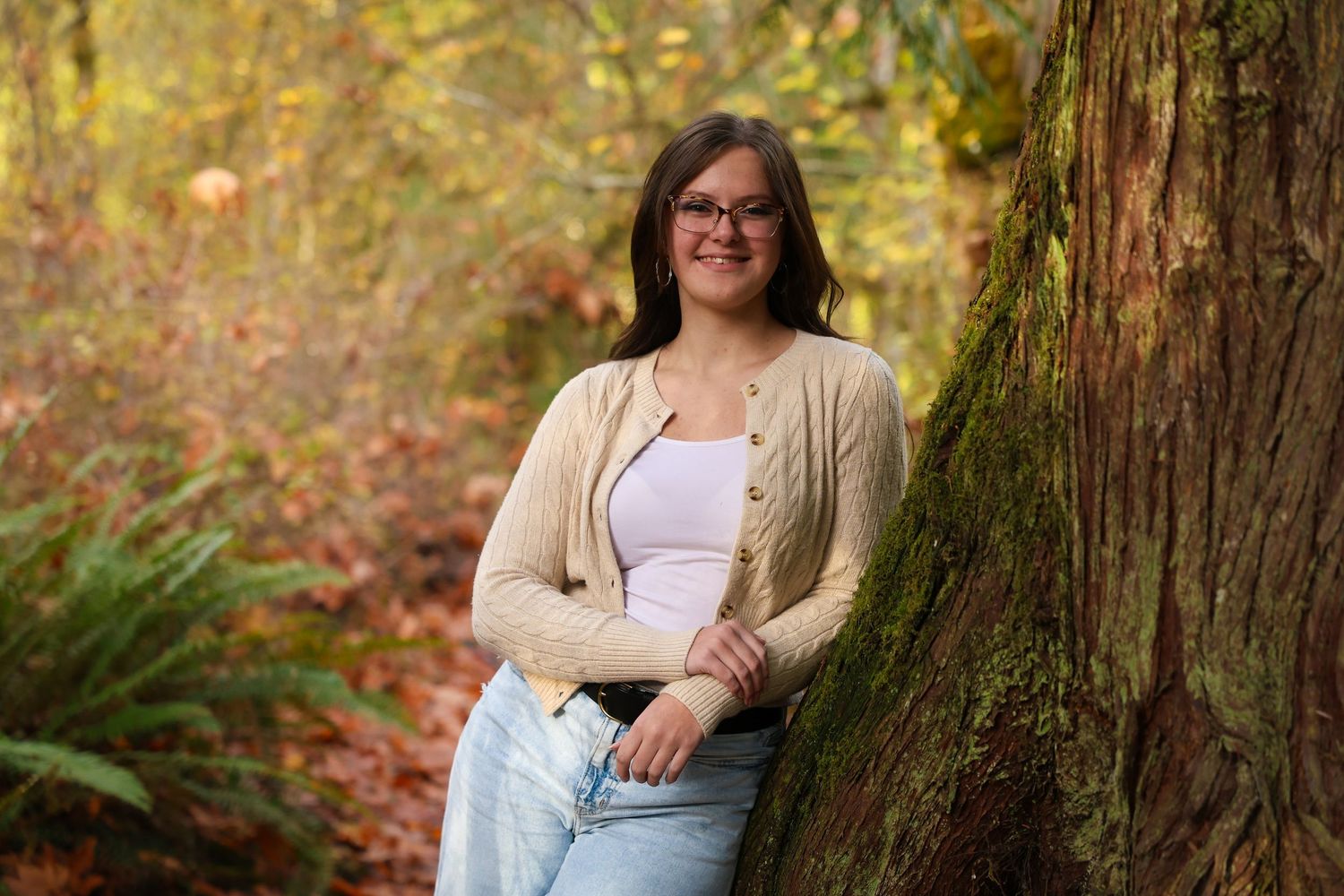 A person in casual attire poses against a mossy tree trunk in a lush forest setting during autumn.