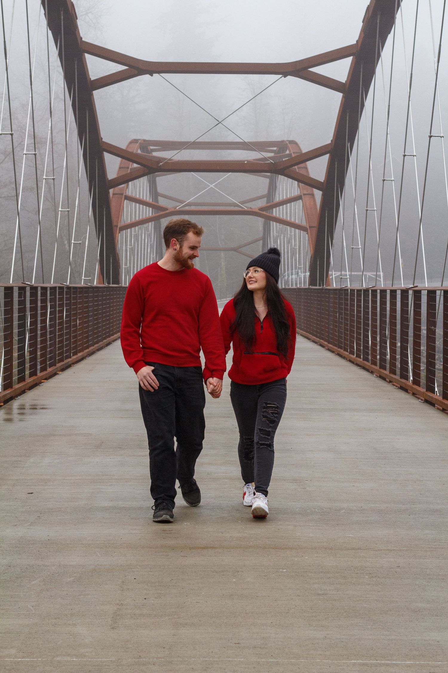 A couple in matching red sweaters walks hand in hand across a foggy bridge.