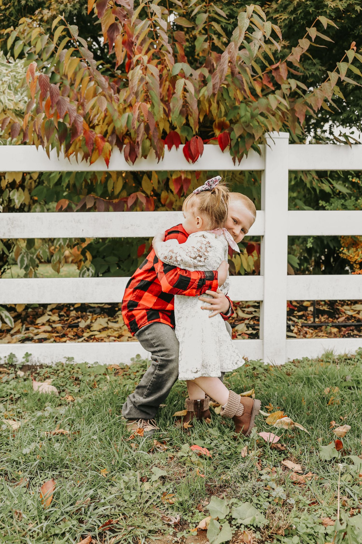 Two young children in autumn outfits play together near a white fence as fall leaves scatter on the grass.