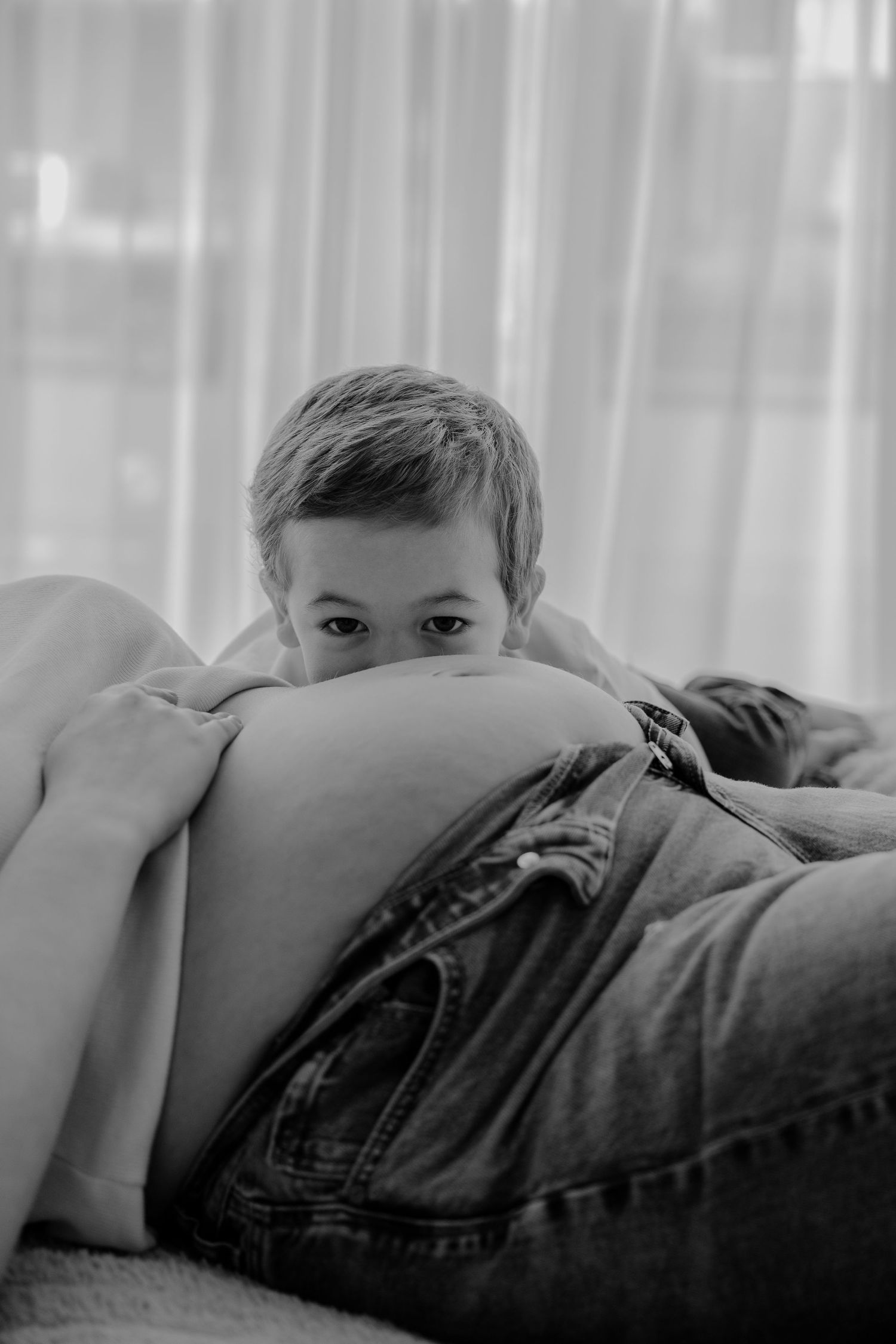 Black and white photo of a young boy peeking over a pregnant woman's bare belly, resting his chin on it.