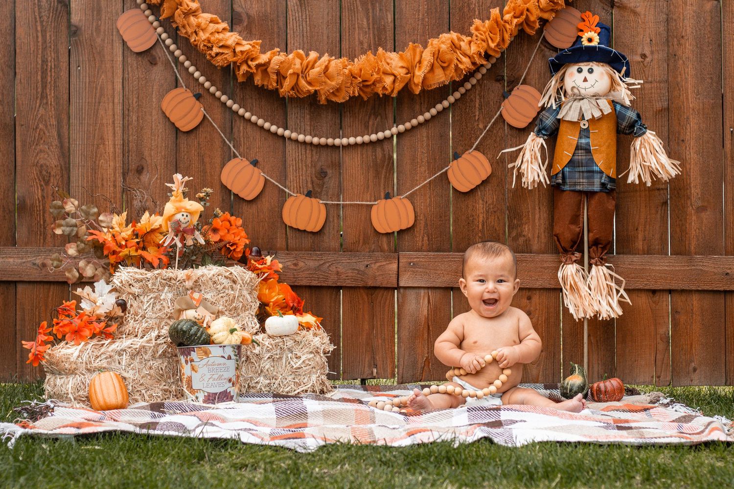 Baby in orange diaper sitting on rustic backdrop with autumn decorations including pumpkins and hay bales.
