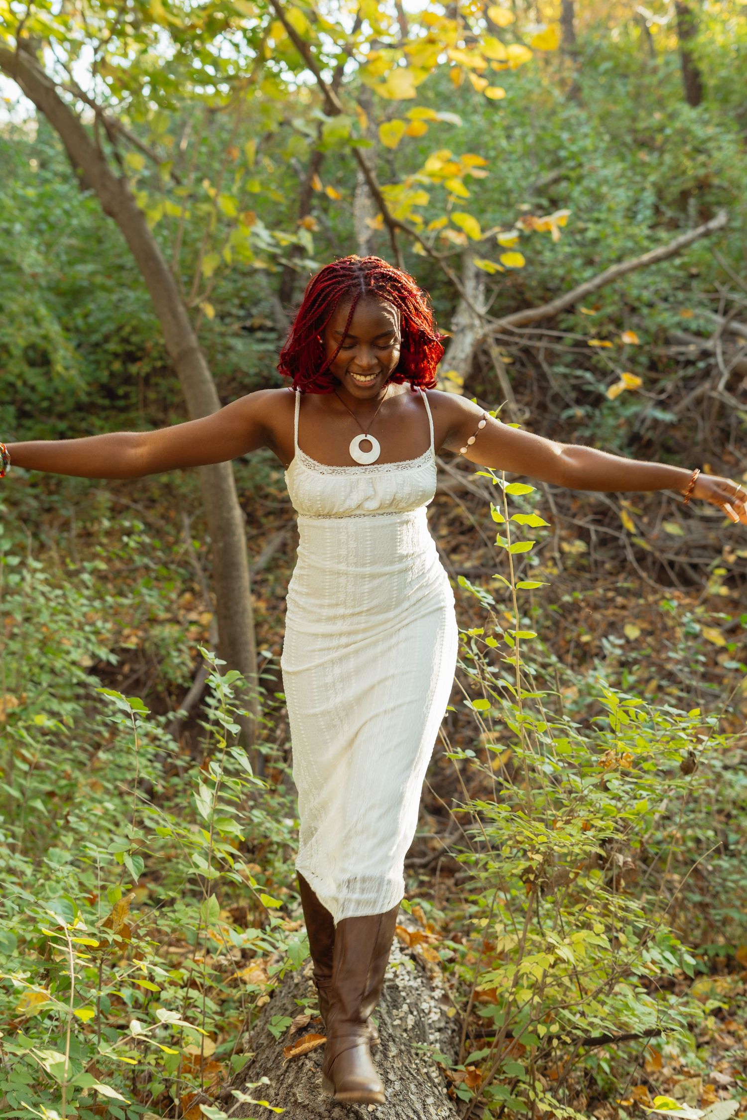 A joyful person in a white dress twirls playfully in a wooded setting with autumn foliage in the background.