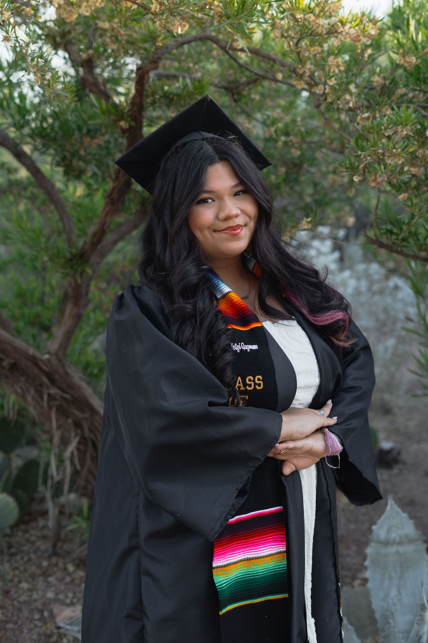 Graduate in black cap and gown with colorful serape stole smiles outdoors among desert plants.