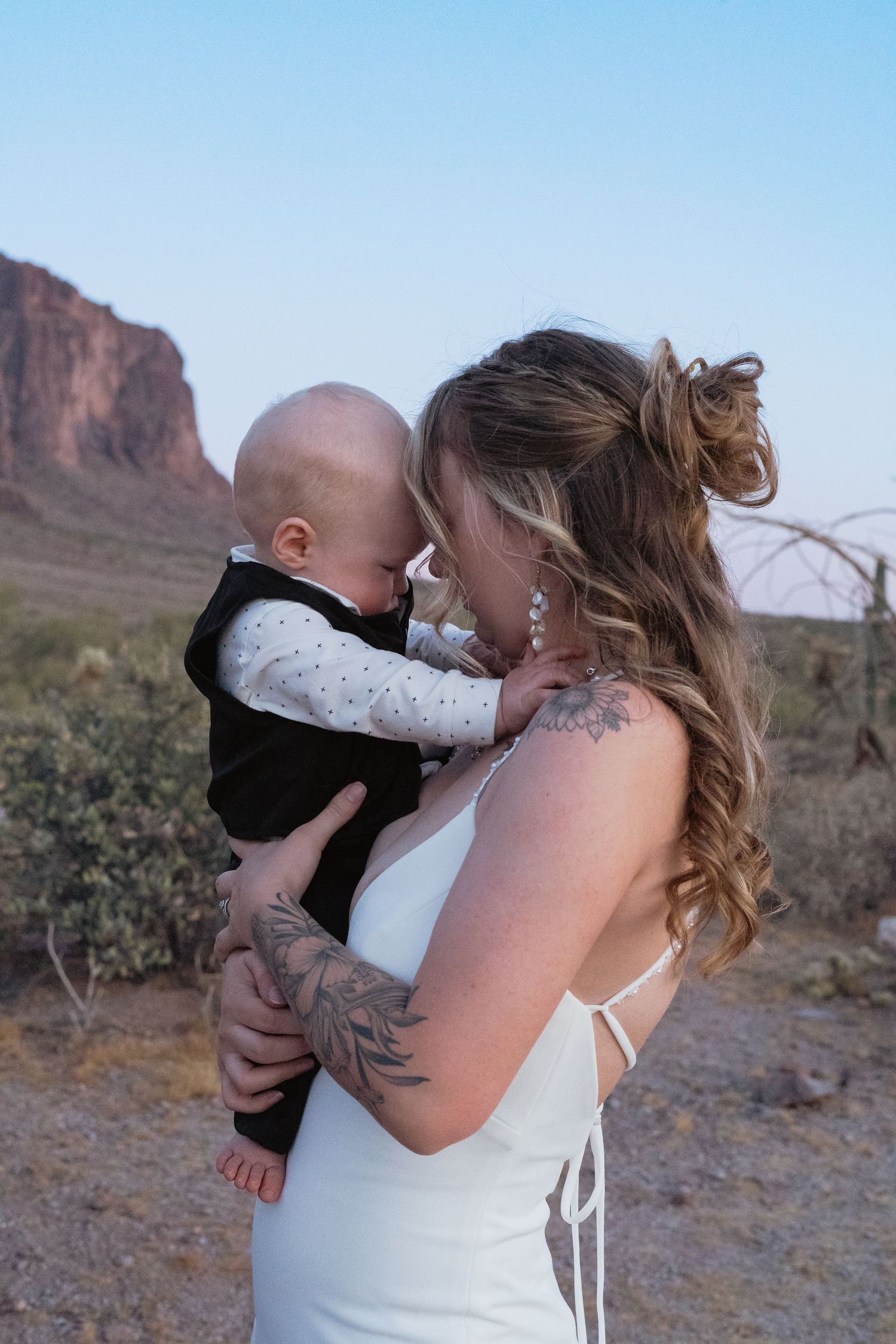 Tattooed bride in white gown tenderly kissing baby while holding them in desert wedding setting.