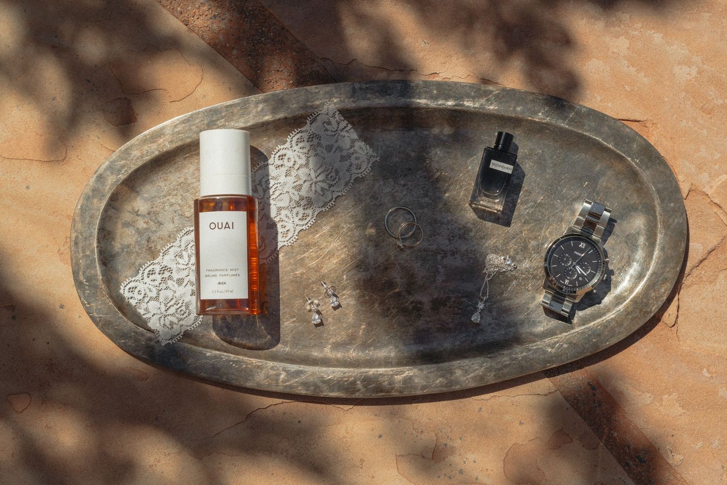 Silver oval tray with Ouai hair oil, dark perfume bottle, watch, and small accessories on a terracotta surface.