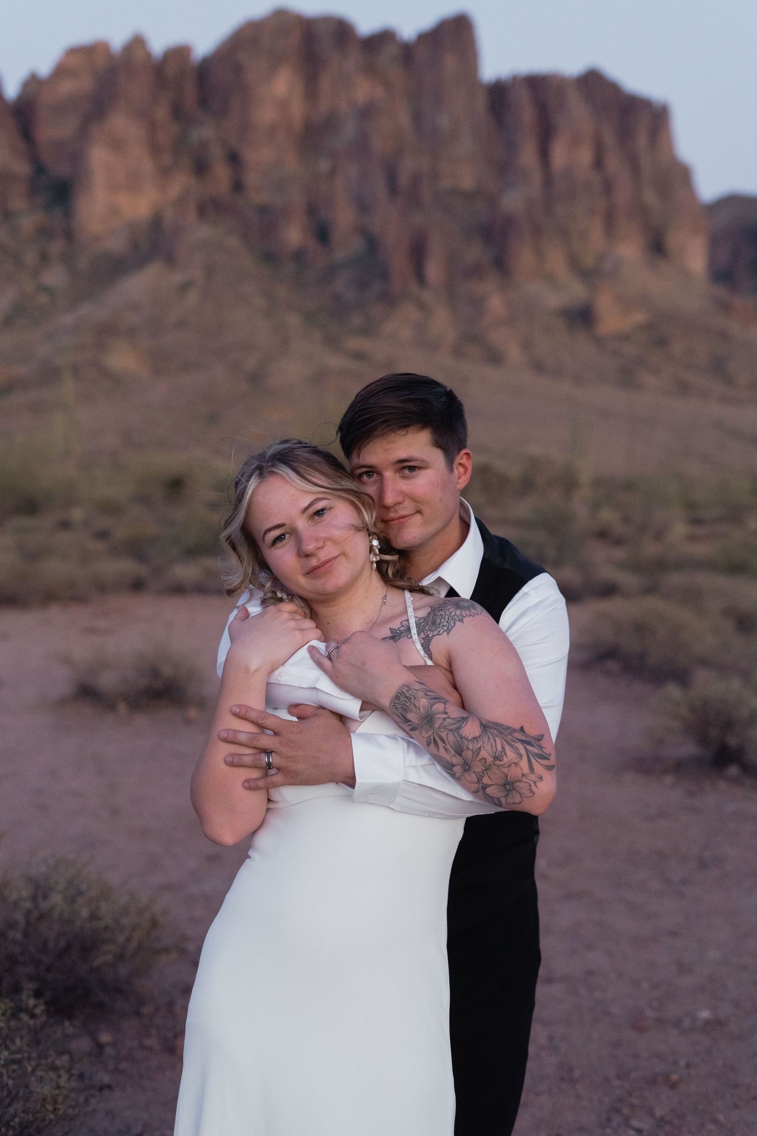 Groom with tattoos embraces bride in white dress with rocky Arizona desert mountains at dusk.
