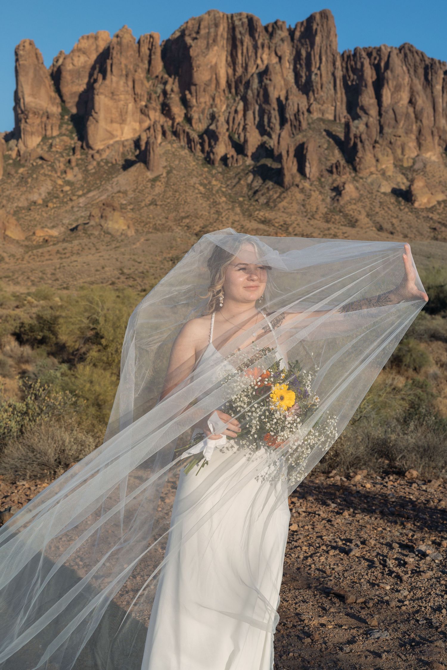 Bride in white gown and flowing veil holds wildflower bouquet with Superstition Mountains backdrop in Arizona desert.