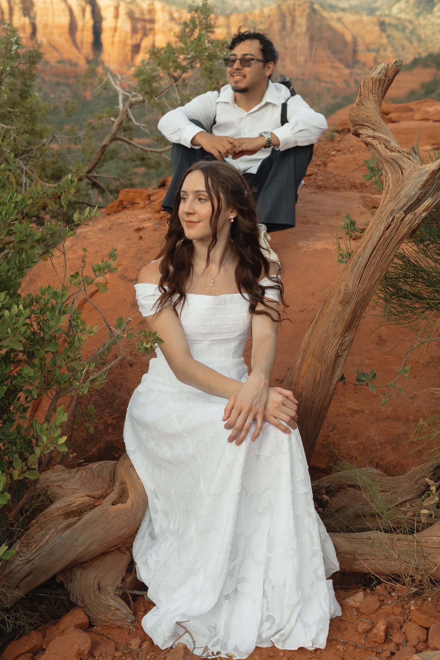 Bride in white gown sits on red rocks in Sedona, AZ, with groom in sunglasses posing behind her.