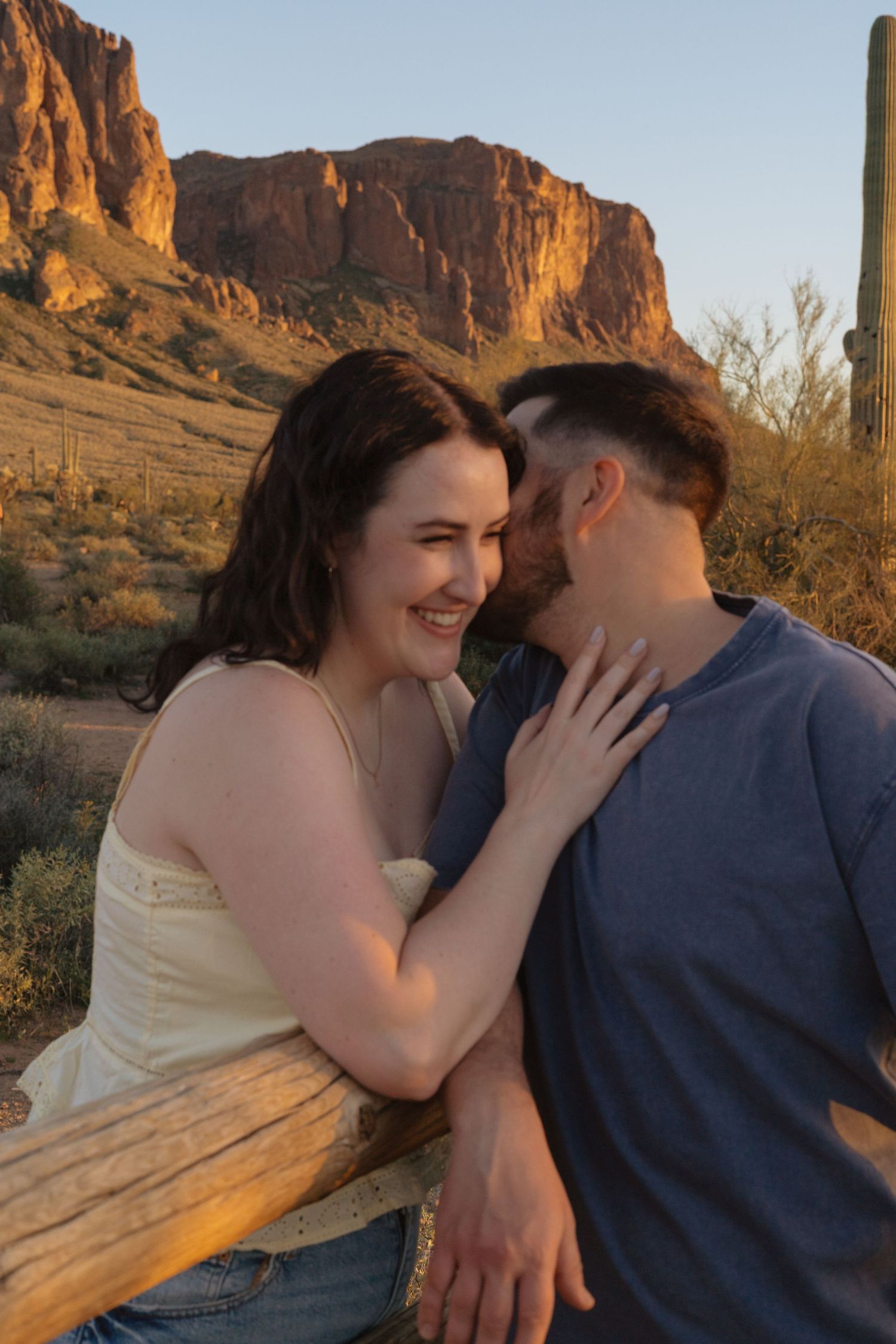 A romantic couple embraces during golden hour with scenic desert mountains and cliffs in the background.