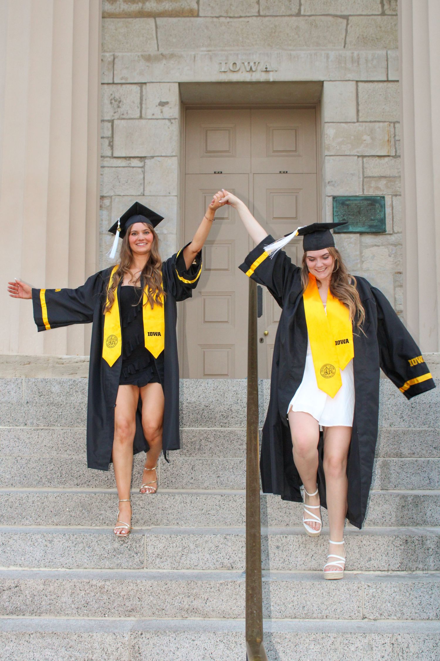 Graduate in cap and gown striking playful poses against stone building columns.