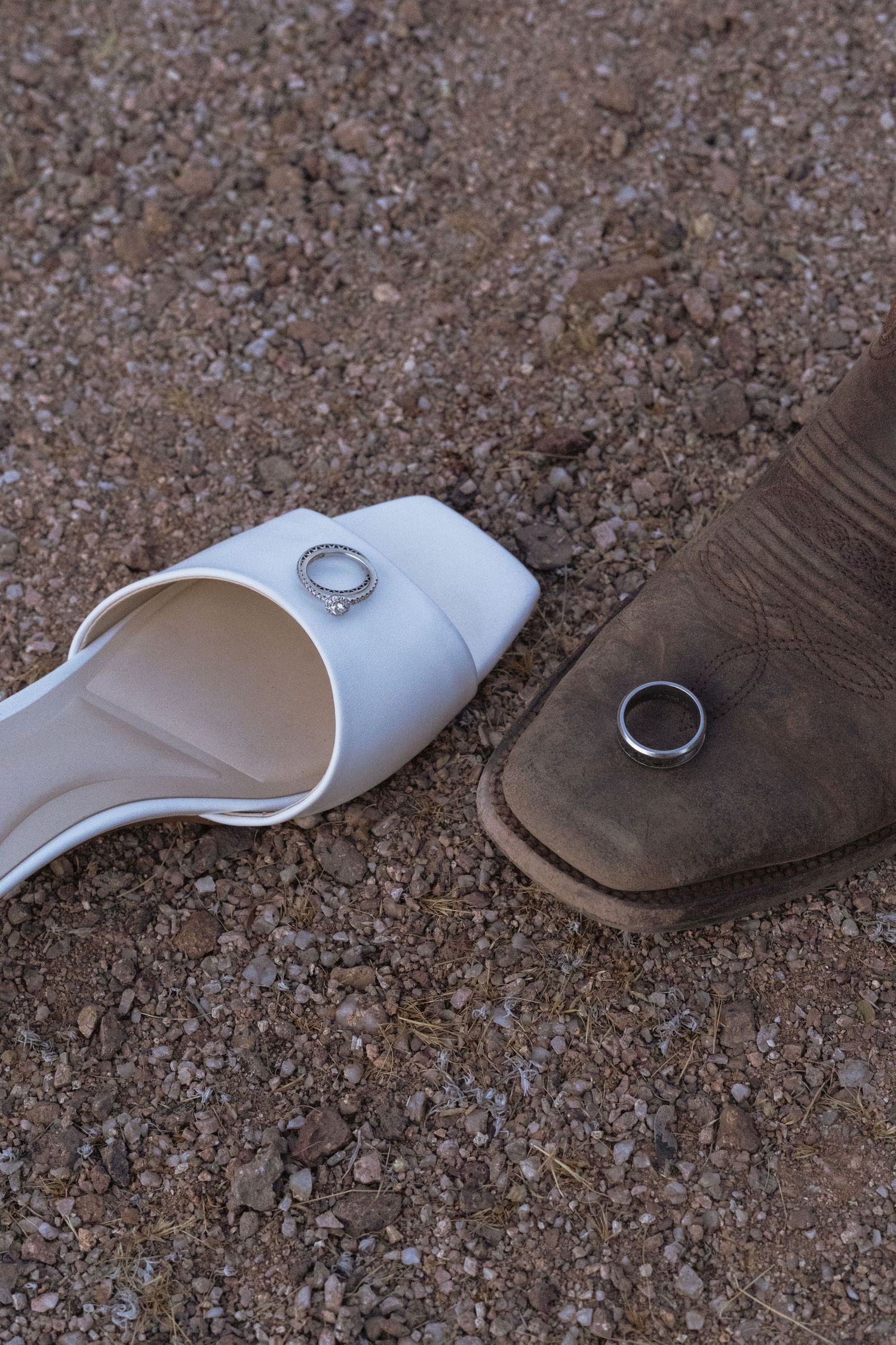 White bridal heel and brown cowboy boot on gravel with wedding rings resting on each shoe.