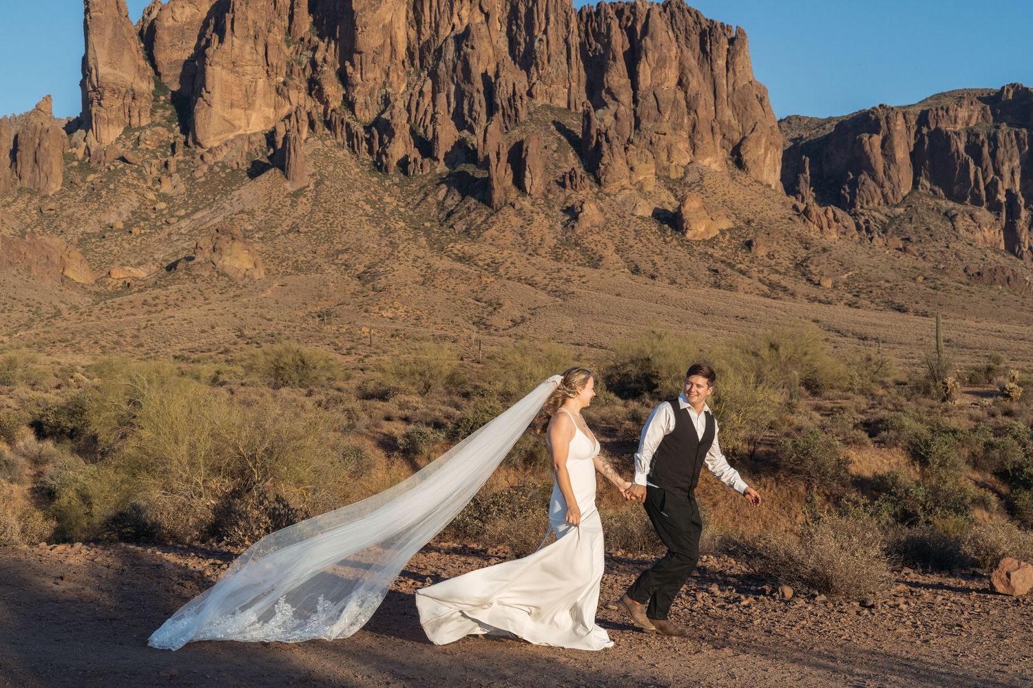Bride and groom walk hand-in-hand in the Arizona desert with Superstition Mountains backdrop, veil flowing in wind.