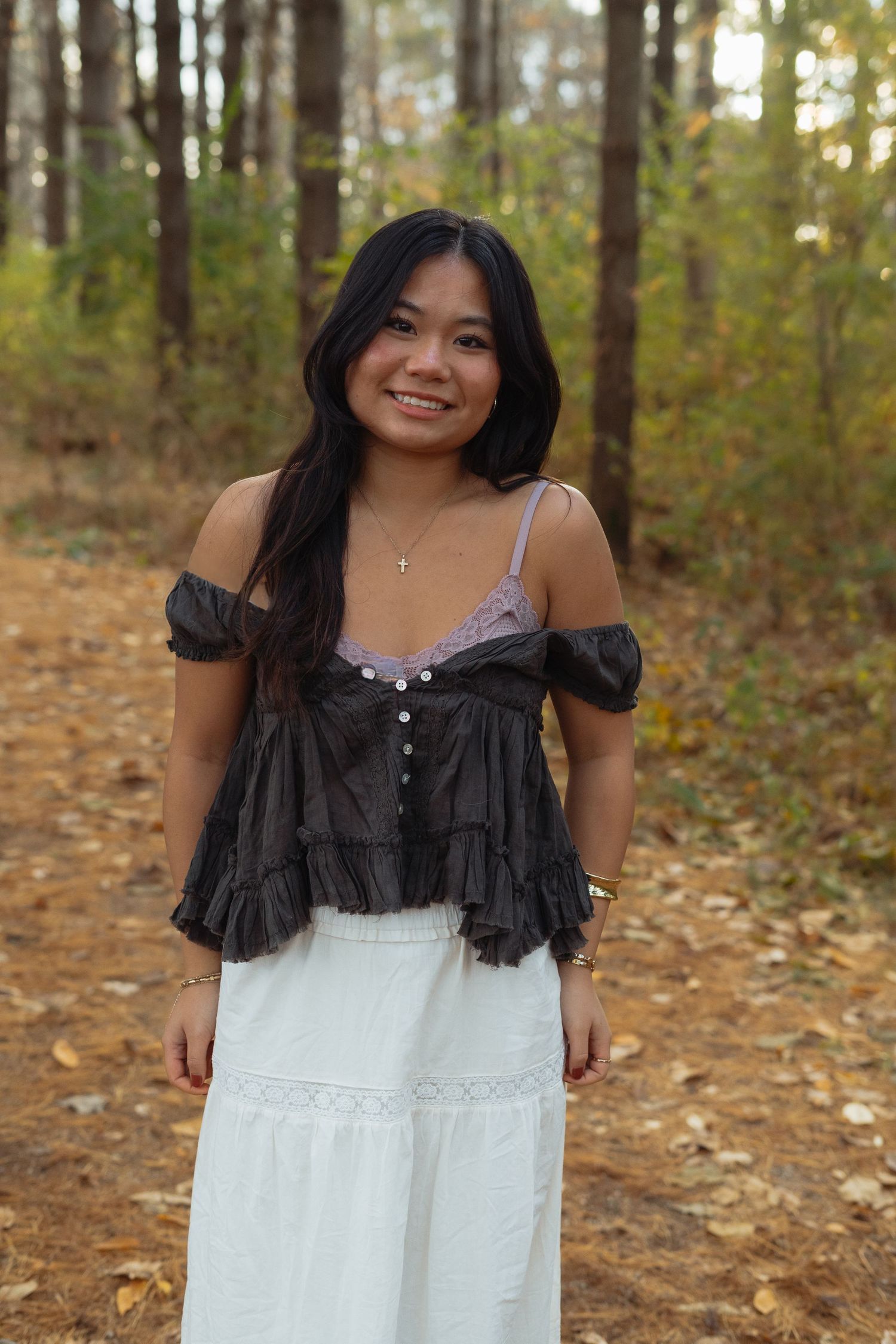 A person wearing a black off-shoulder top and white maxi skirt poses on a leaf-covered trail in an autumn forest.