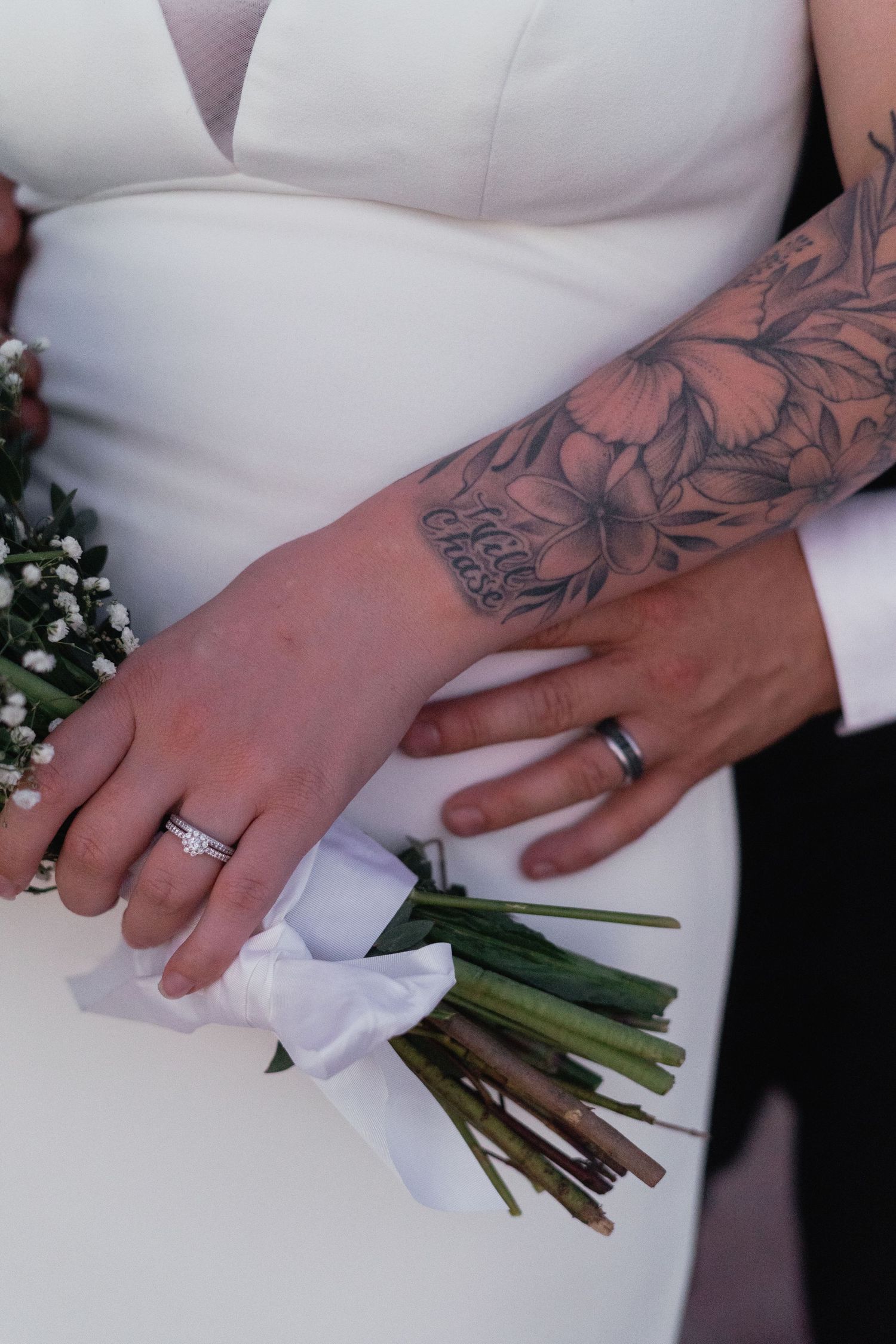 Close-up of bride and groom hands showing wedding rings, bridal bouquet, and groom's floral tattoo sleeve.