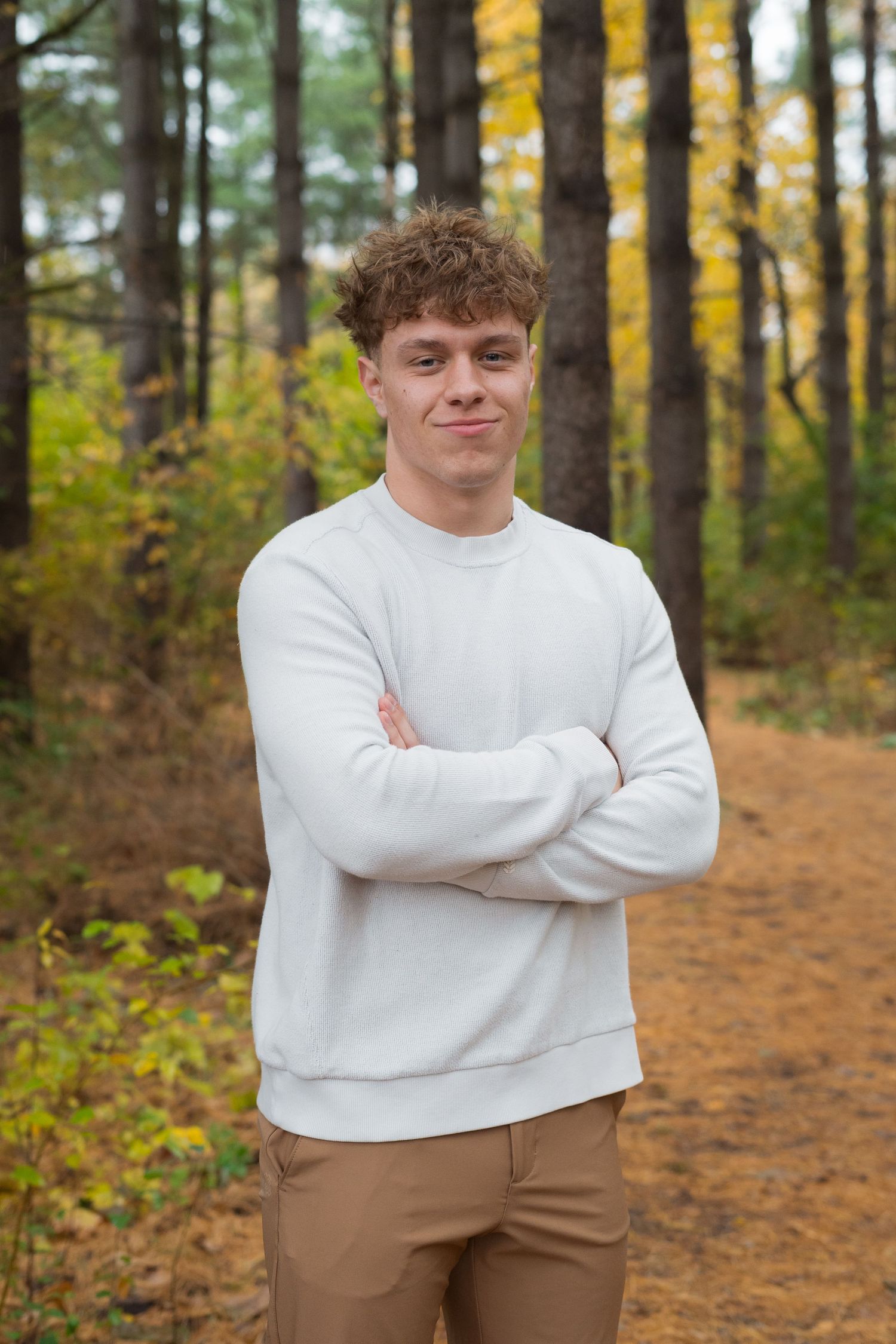 A figure in a white sweater and khaki pants stands on a forest path surrounded by autumn trees and fallen leaves.