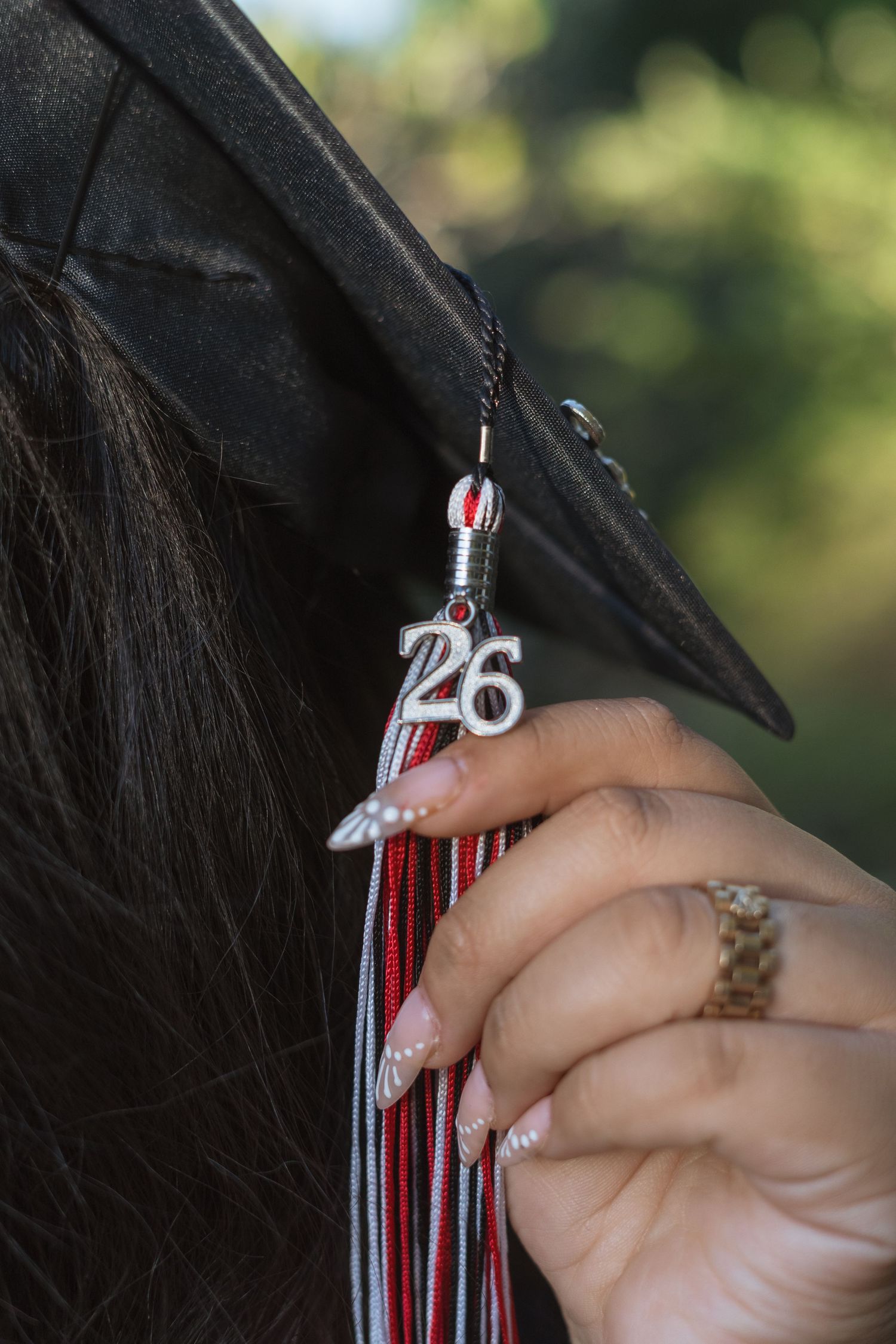 Close-up of a graduation cap tassel with a silver 26 charm, held by a hand with decorated nails.