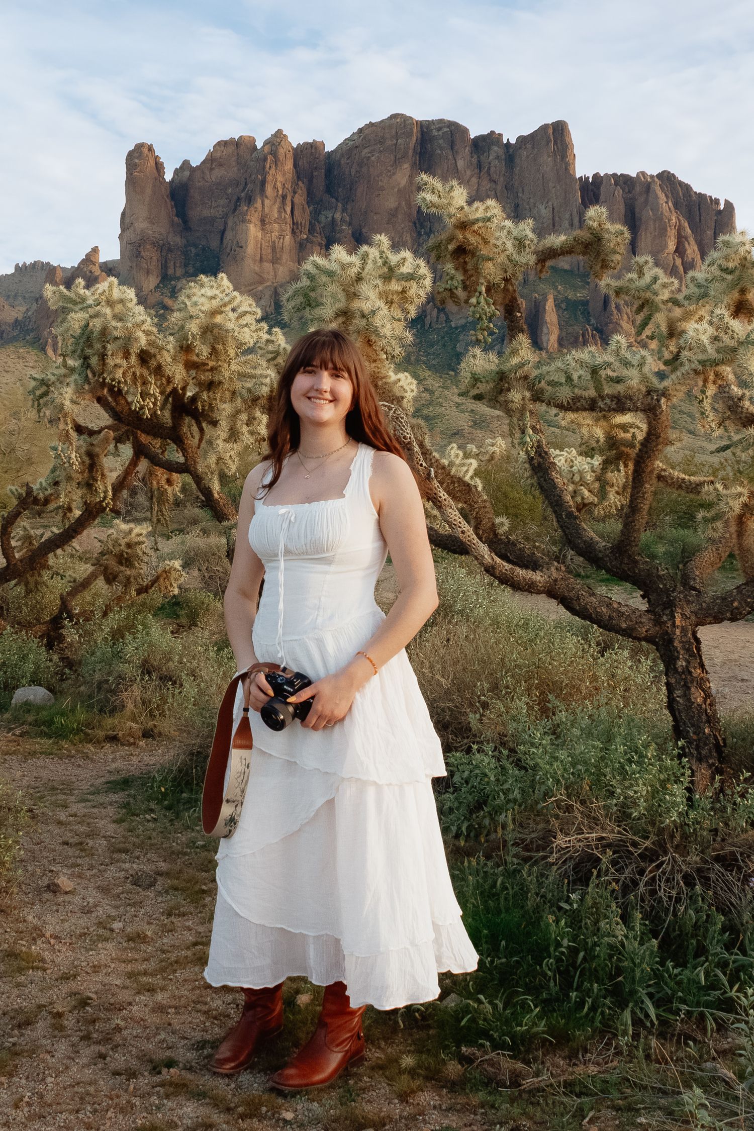 A desert landscape features a person in a white dress and boots standing among cholla cactus with red rock formations.