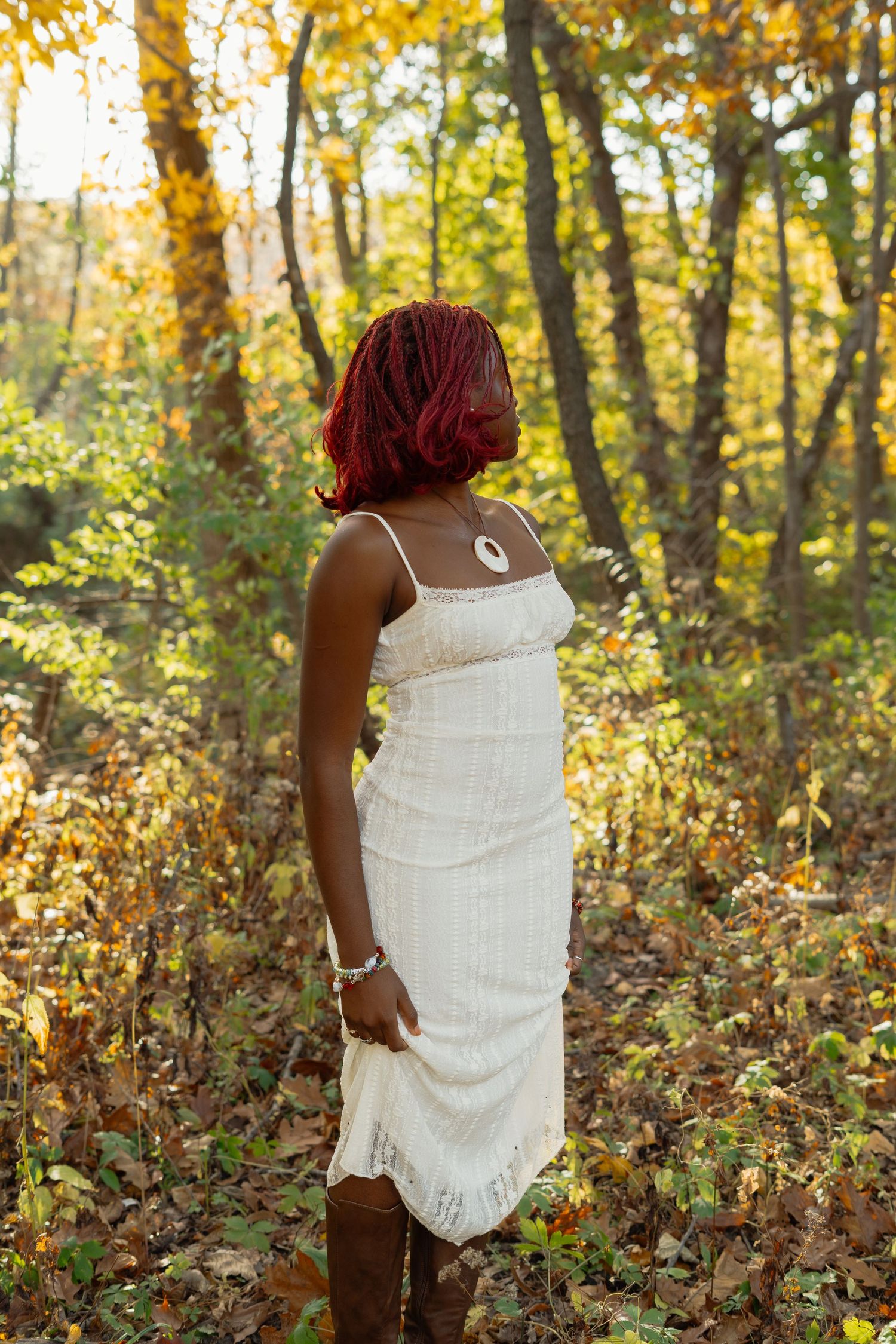 A person in a white knit sweater sits among autumn foliage in a sunlit forest setting.