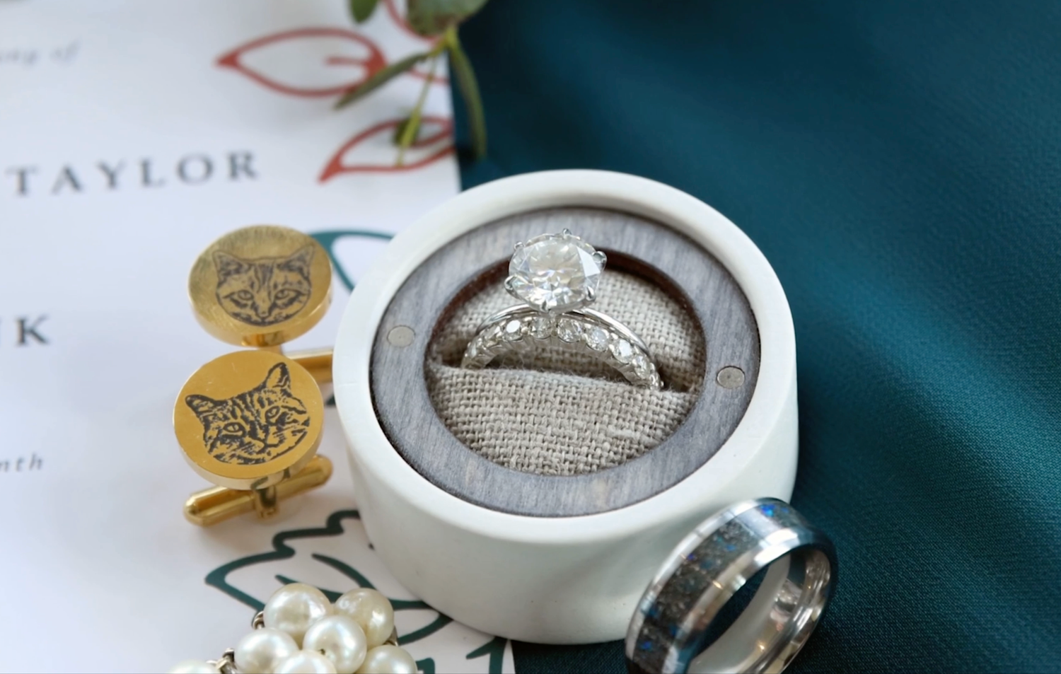 Wedding rings displayed in a circular concrete holder next to gold wax seals and white flowers on teal background.
