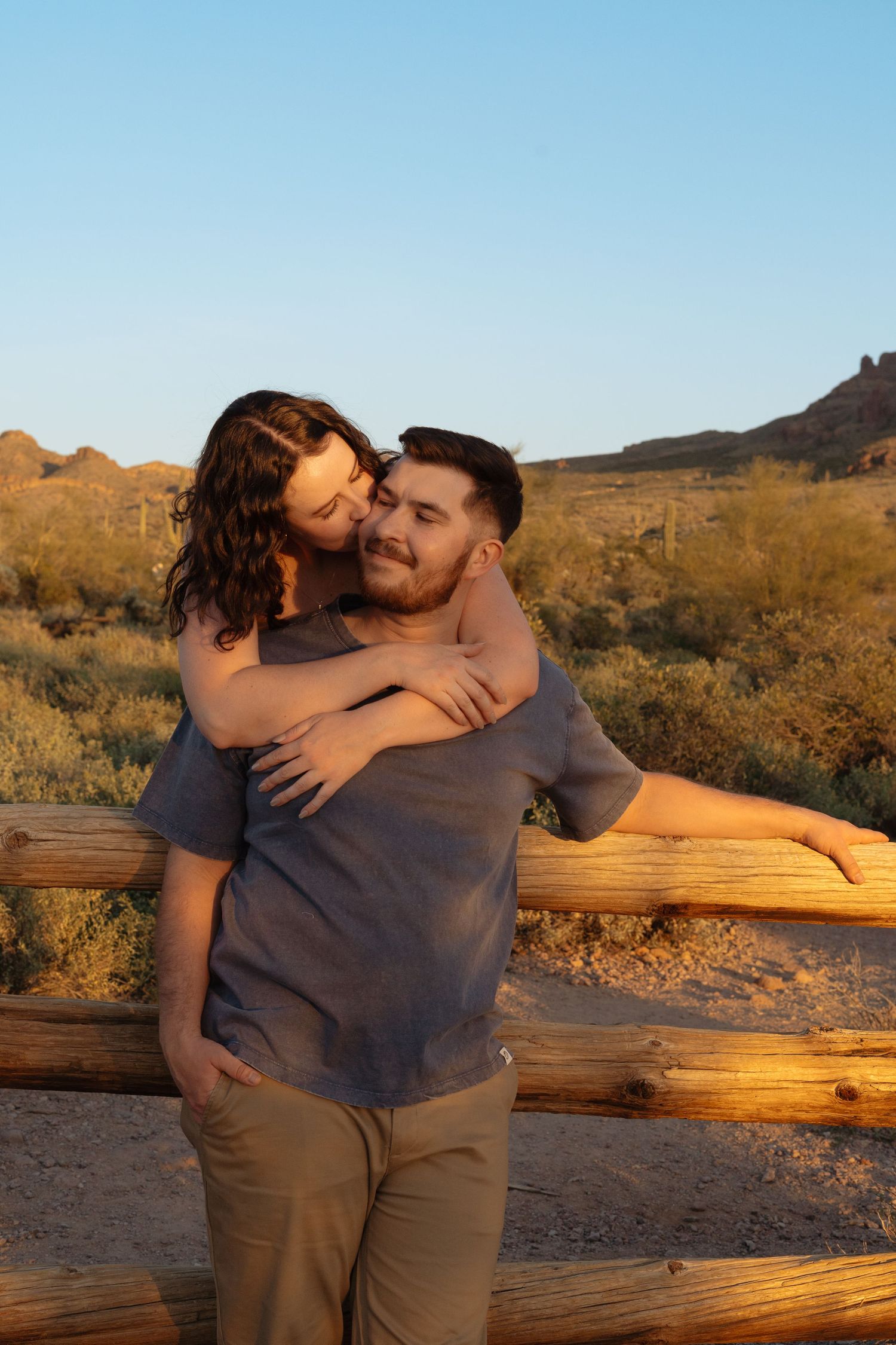 A couple embraces at sunset in front of a wooden fence and desert landscape with mountains in the background.