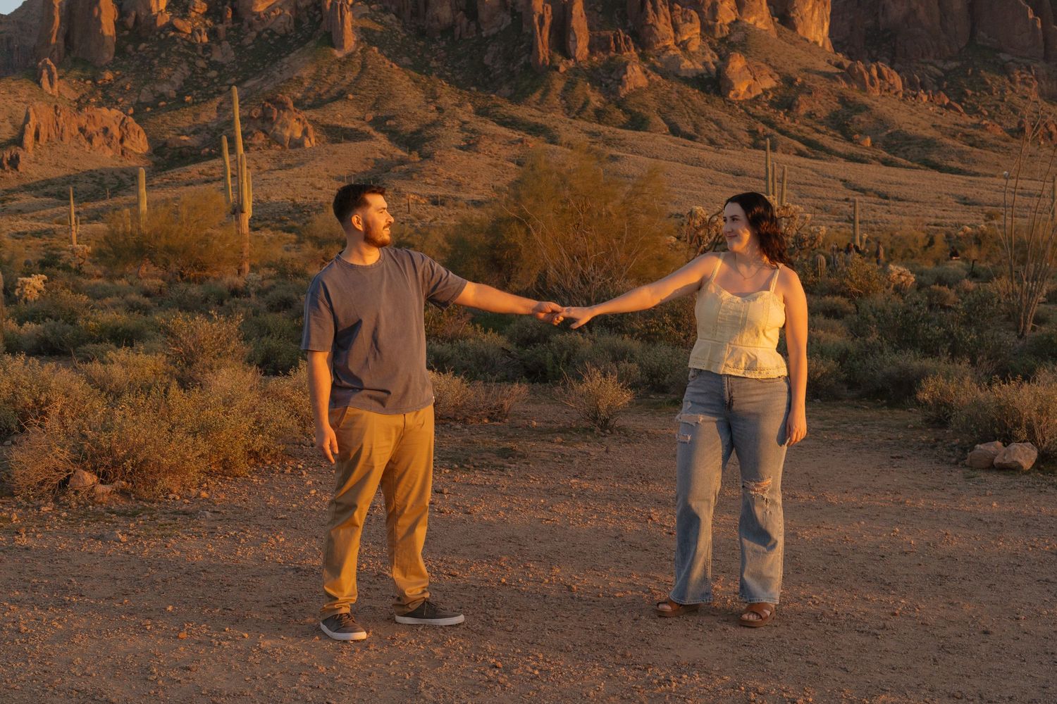 People walking on a desert trail at sunset with warm golden lighting casting long shadows across the sandy terrain.