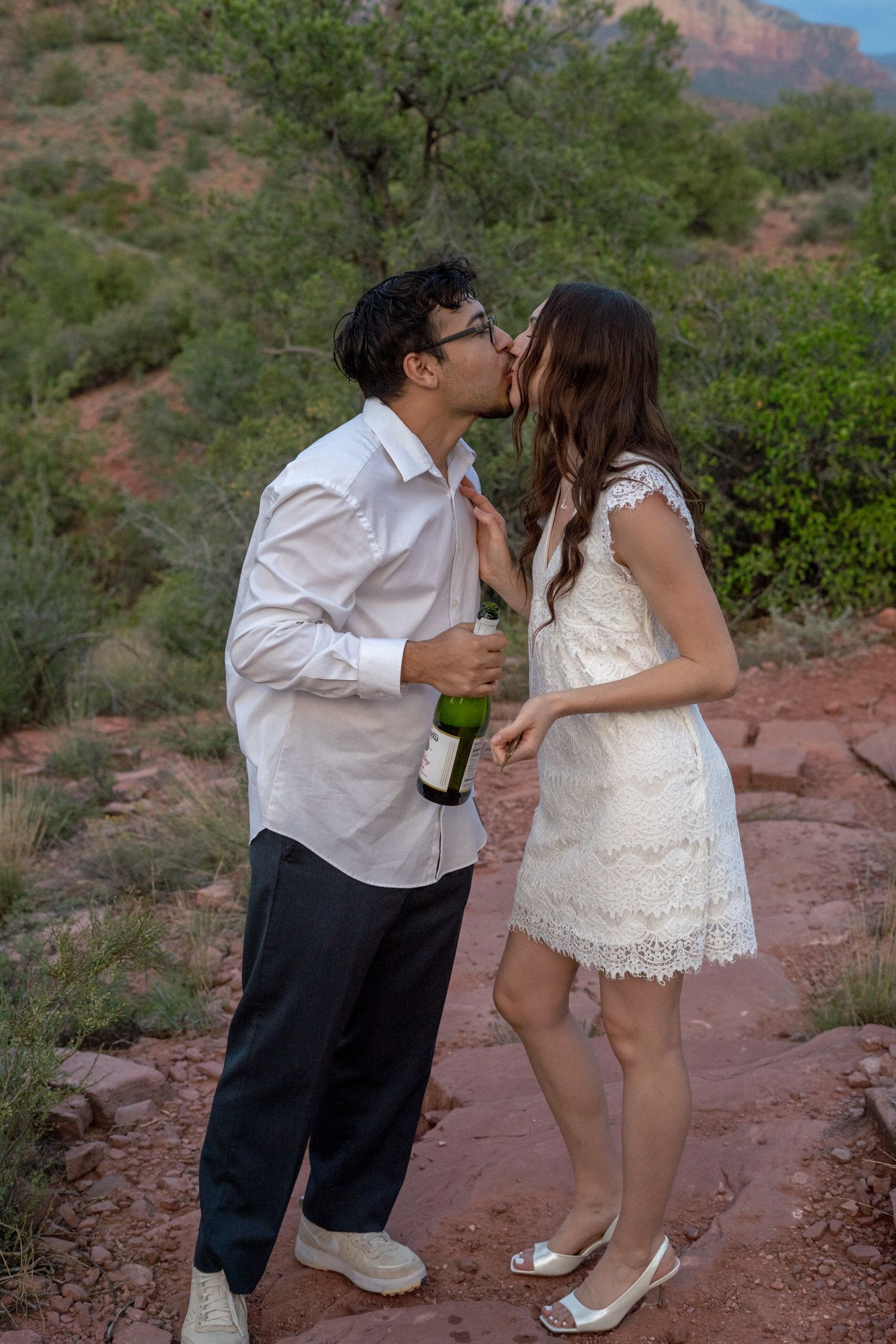 Couple sharing a romantic kiss outdoors in Sedona, Arizona, man holding champagne bottle, woman in white lace dress.