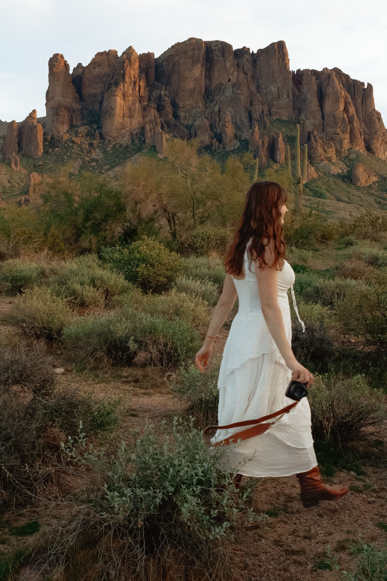 A person in a white dress walks along a desert trail with dramatic red rock formations of the Superstition Mountains in view.