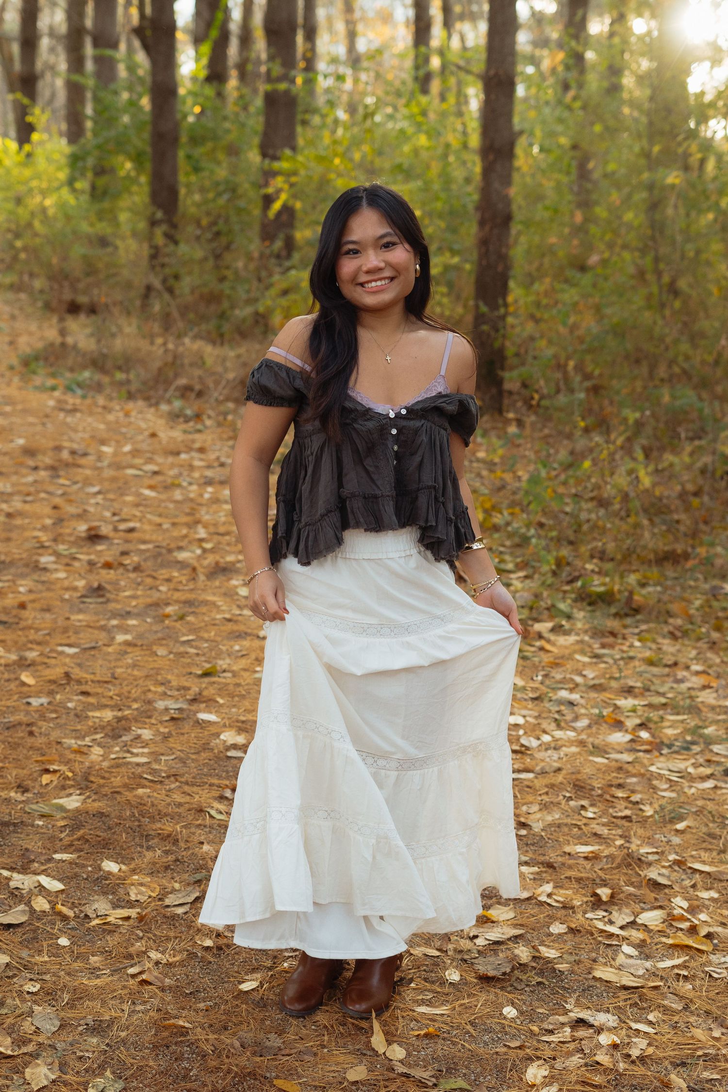 A person in a white dress sits gracefully among autumn leaves in a wooded setting during golden hour.