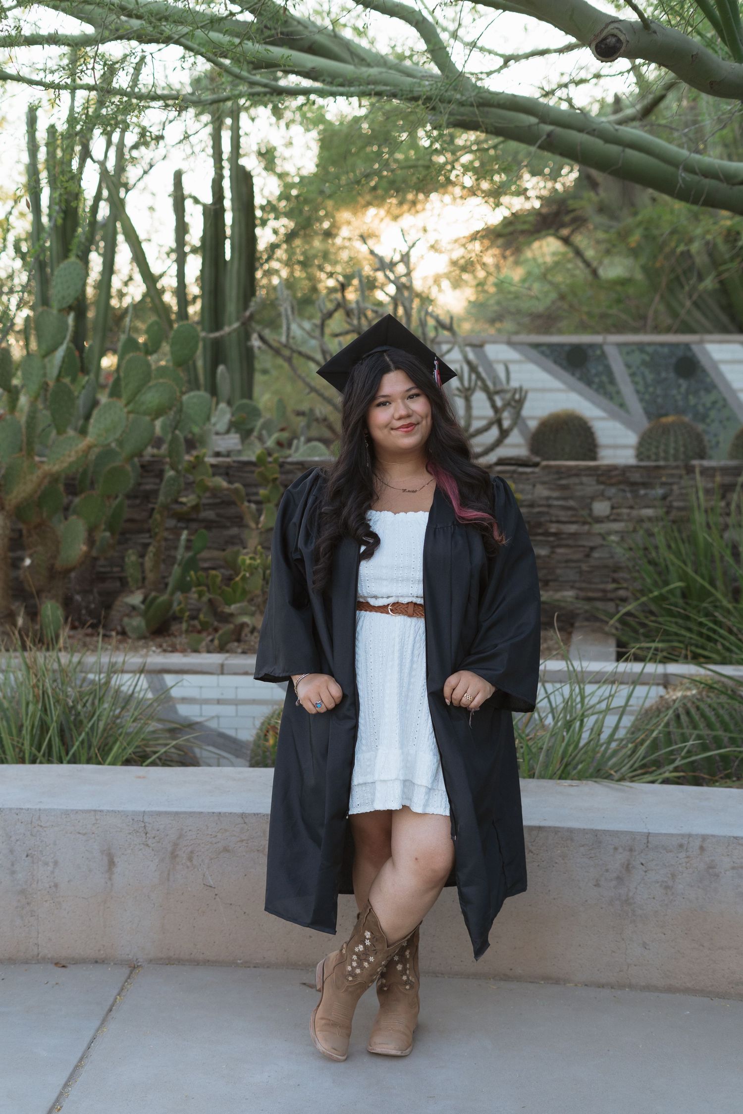 Young woman in graduation cap and gown over white dress poses outdoors among desert cacti at golden hour.