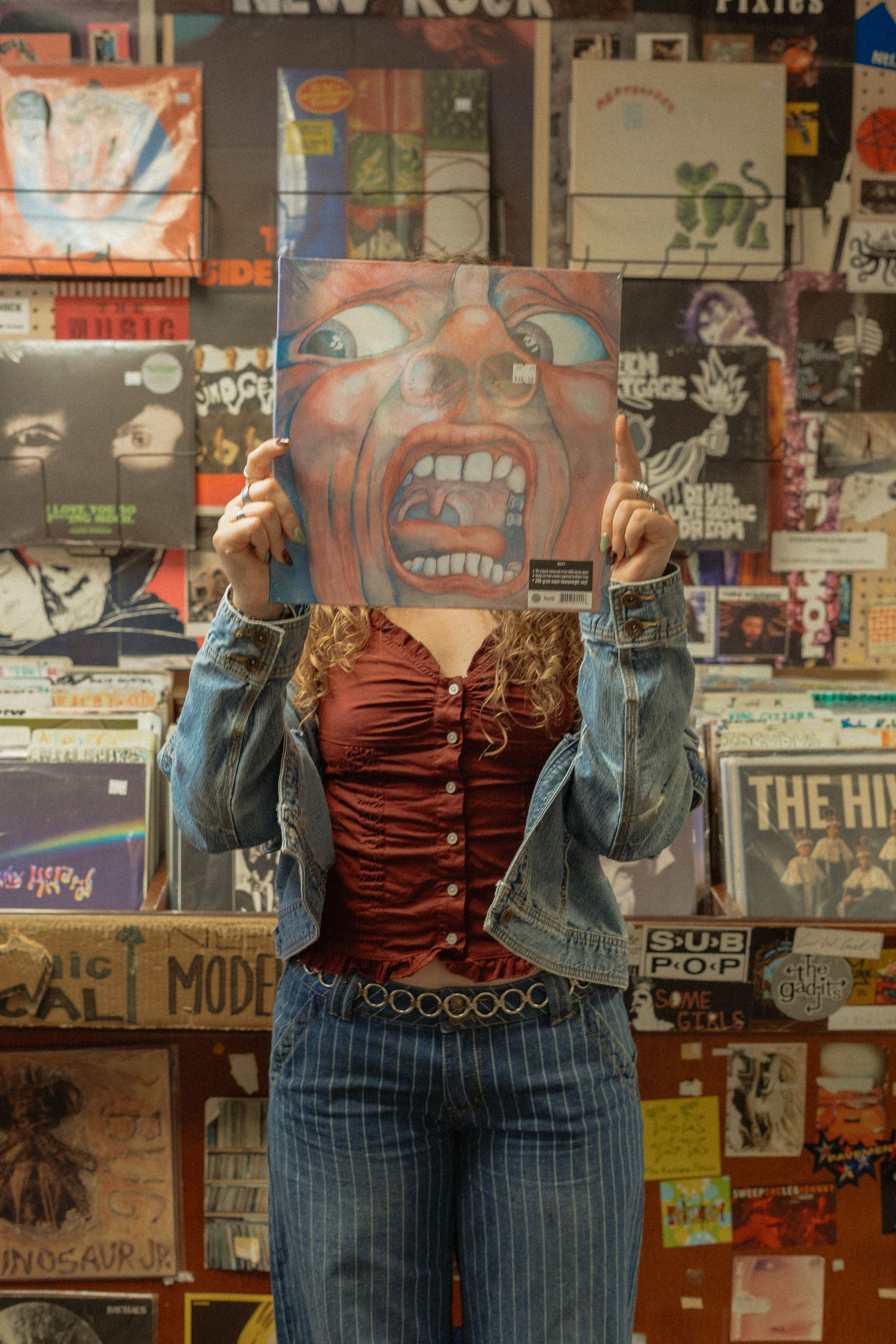 Vintage clothing display showing a red corset top paired with pinstriped jeans against a wall covered in music posters.