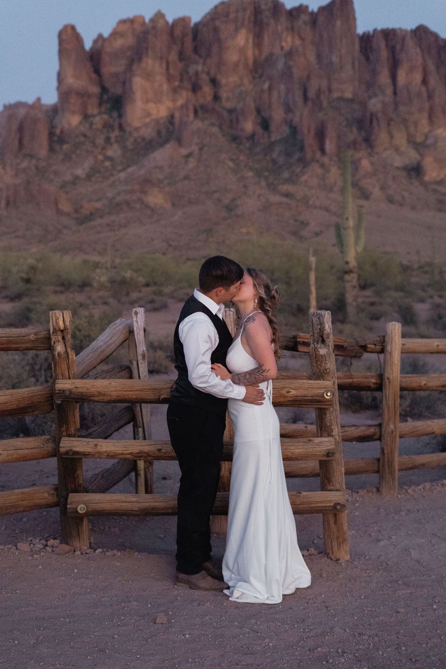 Couple sharing first kiss in wedding attire before rustic fence with Superstition Mountains backdrop at sunset.