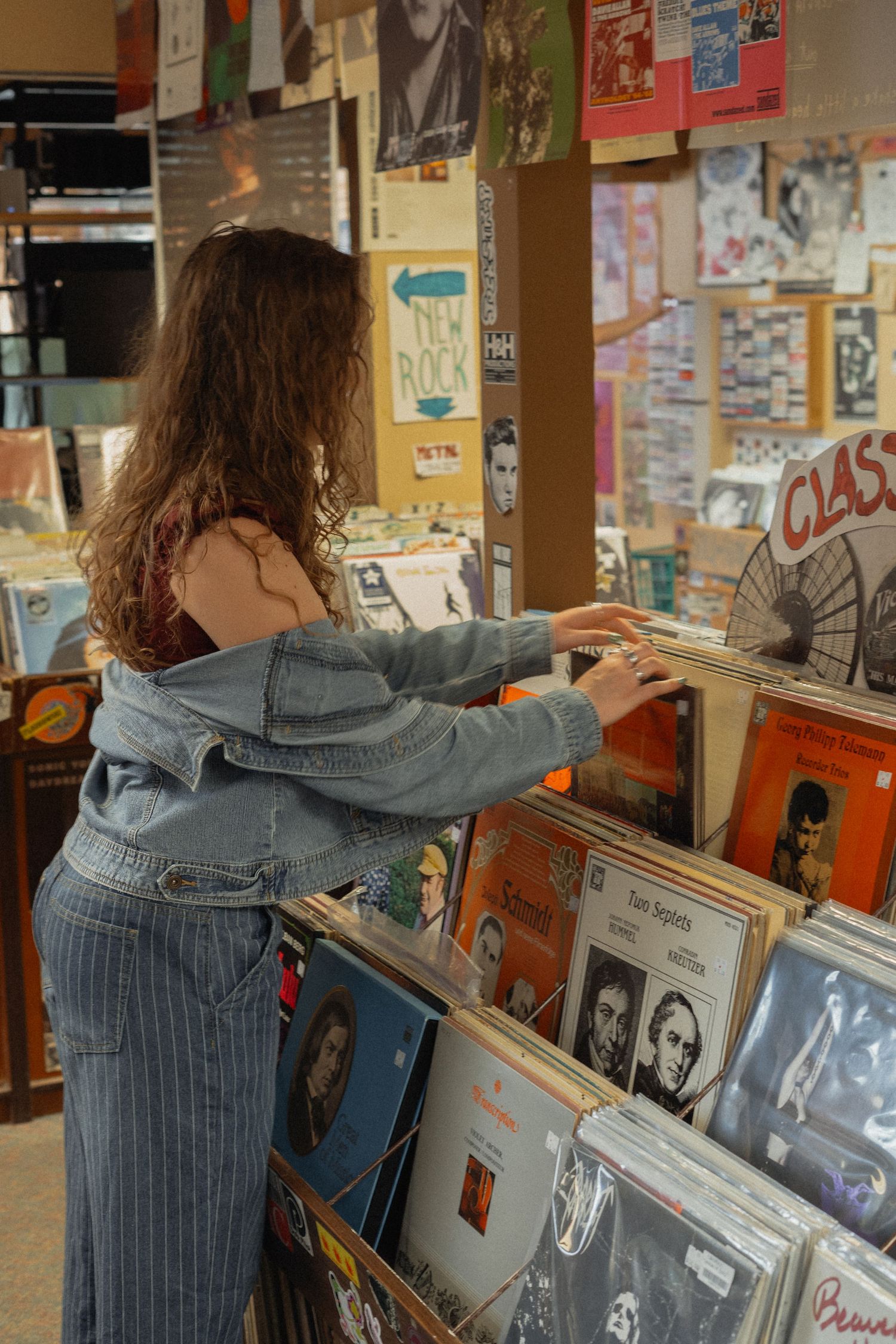 Person in denim jacket browsing through vinyl records at a music store with posters on the walls.