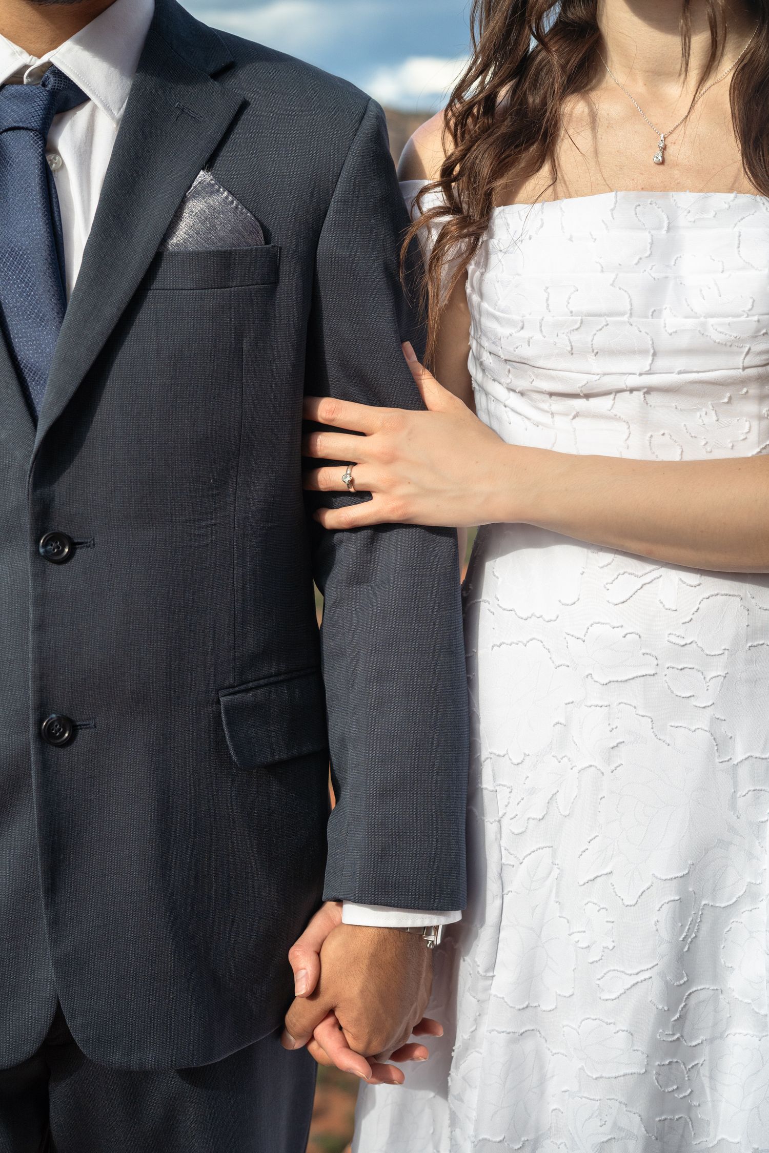 Bride in white strapless dress holds groom's hand in gray suit, showing engagement ring at outdoor wedding.