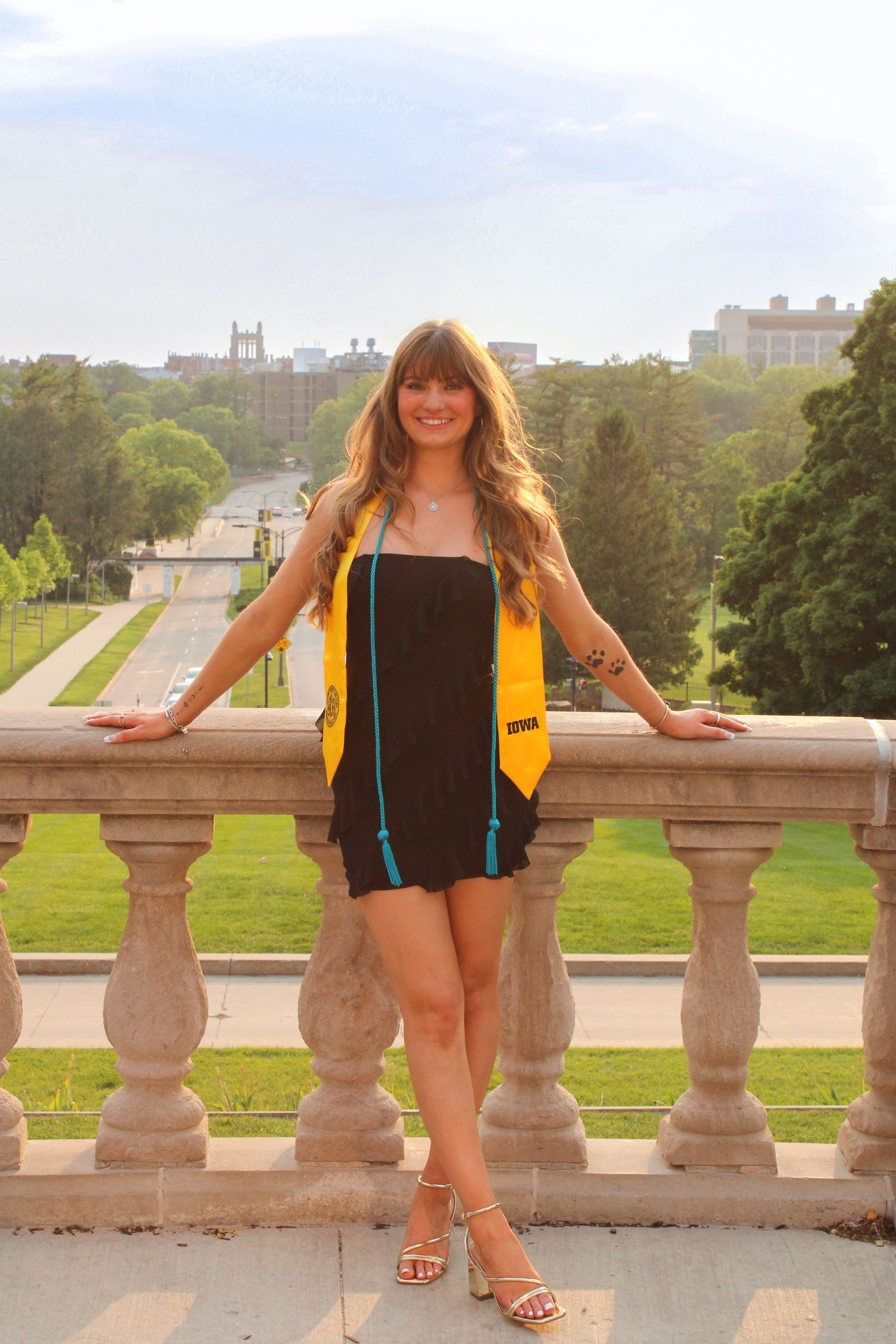A graduate in a black dress and yellow sash poses on a balcony overlooking a scenic campus view.