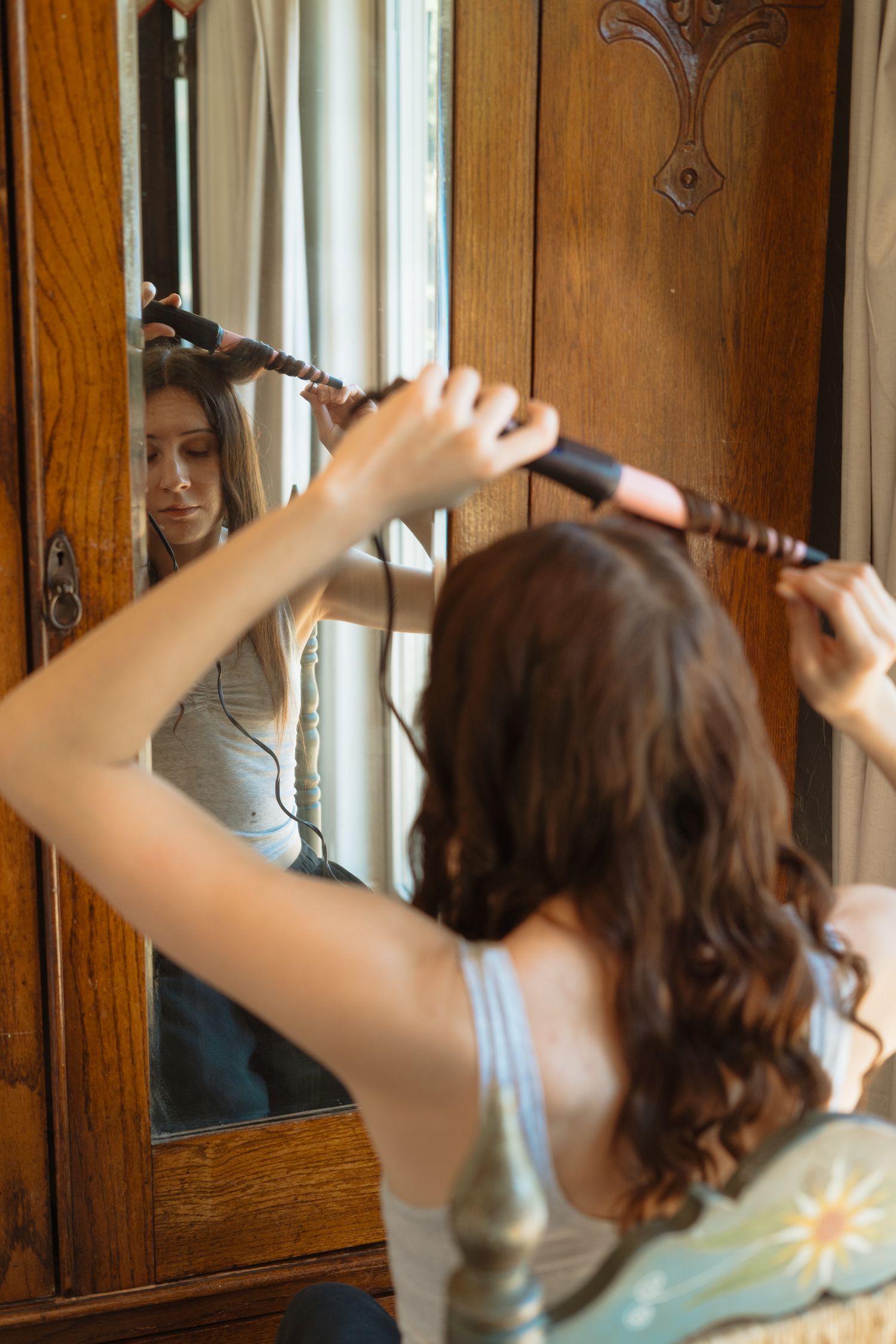 Young woman curling her long brown hair with a curling iron while looking in a vintage wooden mirror.