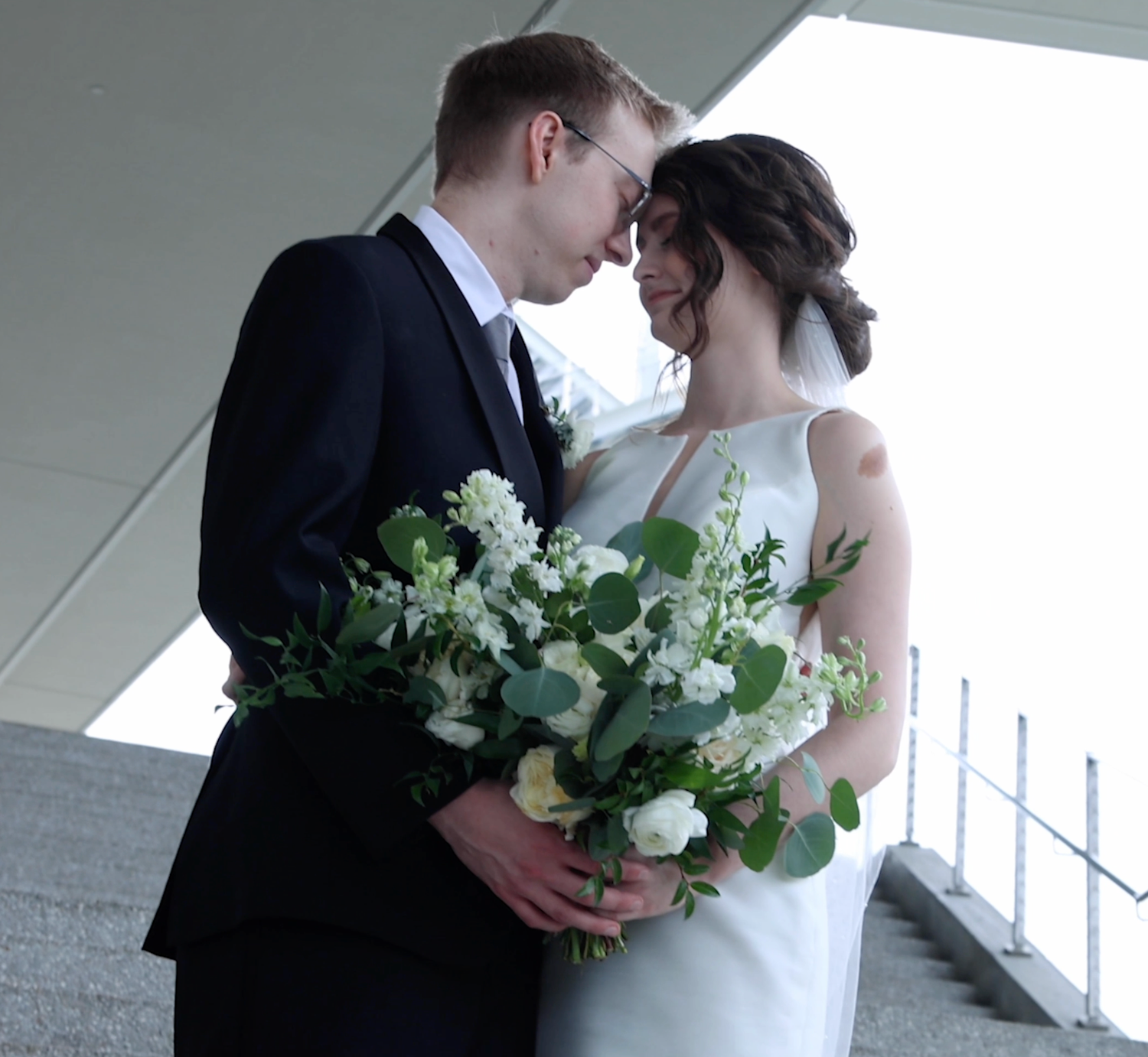 A romantic wedding couple embraces in a modern building with a white floral bouquet.