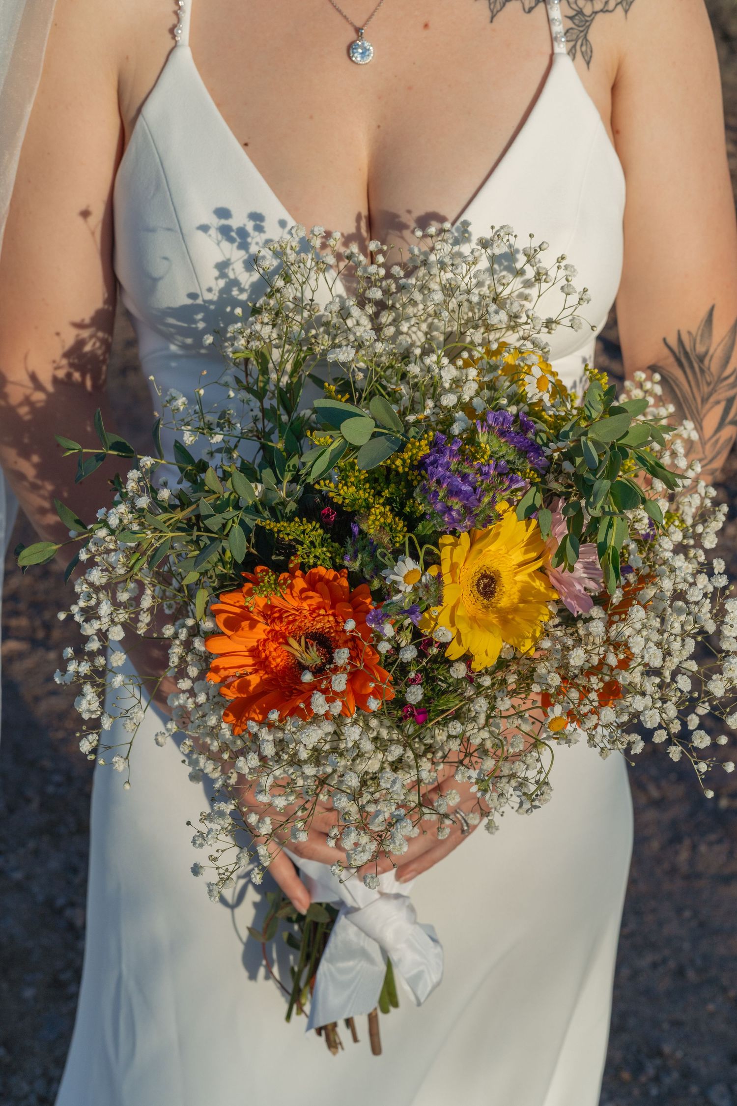 Close-up of colorful wildflower bridal bouquet with gerbera daisies and baby's breath held by tattooed bride in white dress.