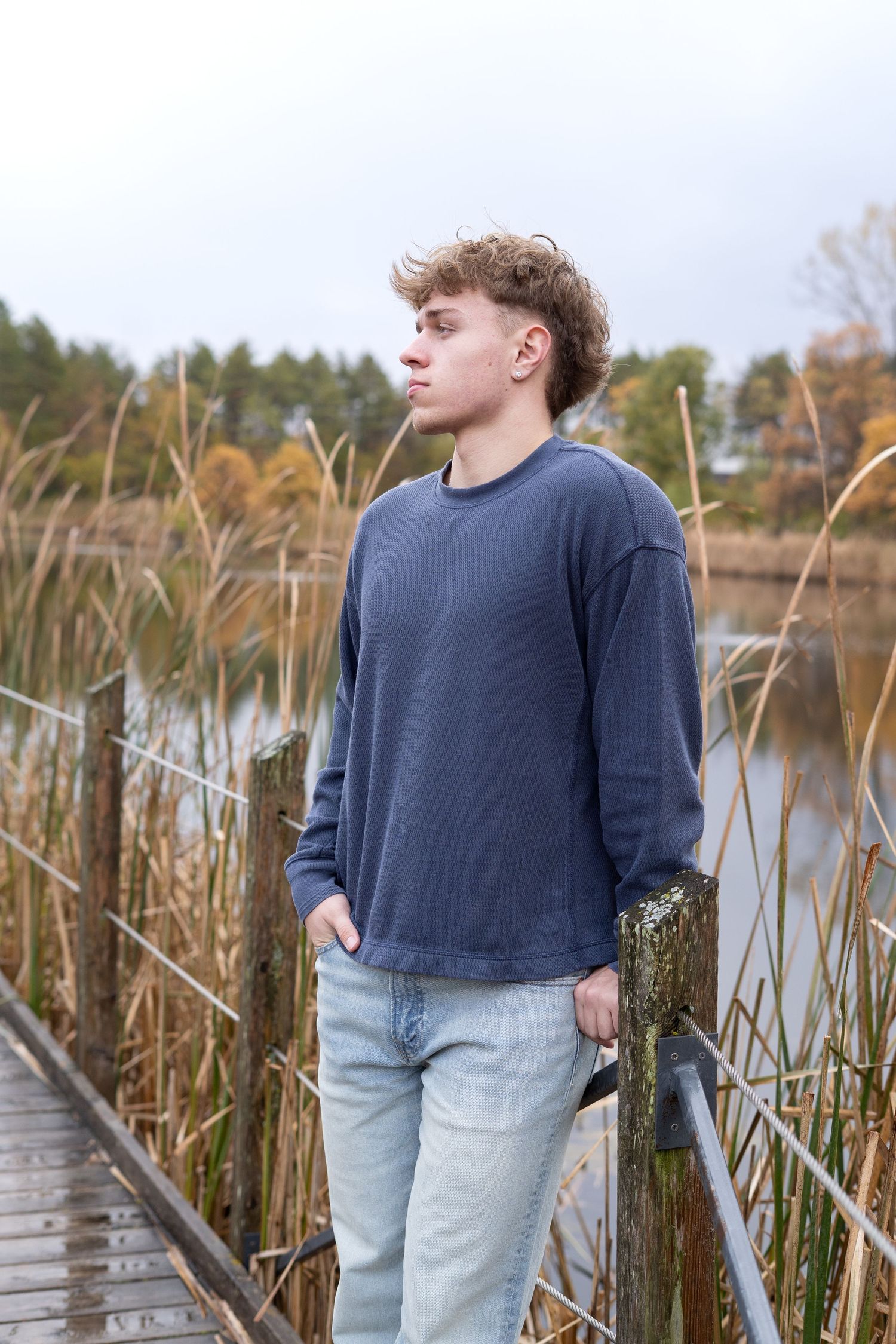 Person in navy sweater and light jeans standing on wooden boardwalk by lake with tall reeds at sunset.
